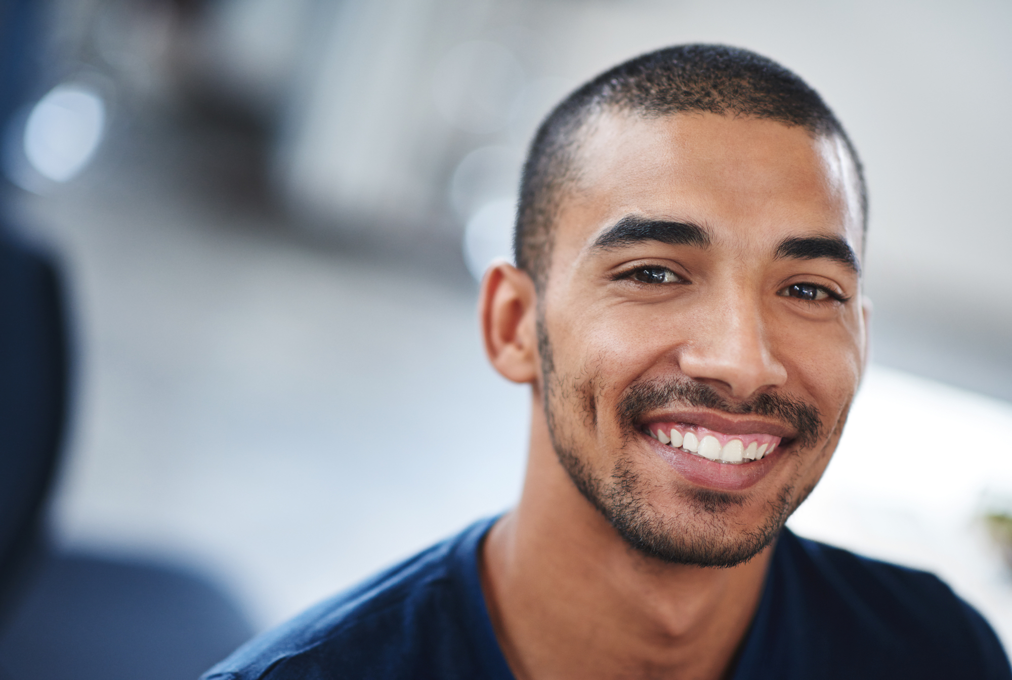 A young man with short hair and a beard smiles warmly at the camera. He is wearing a dark blue shirt, and the background appears to be softly blurred, focusing the attention on his cheerful expression.