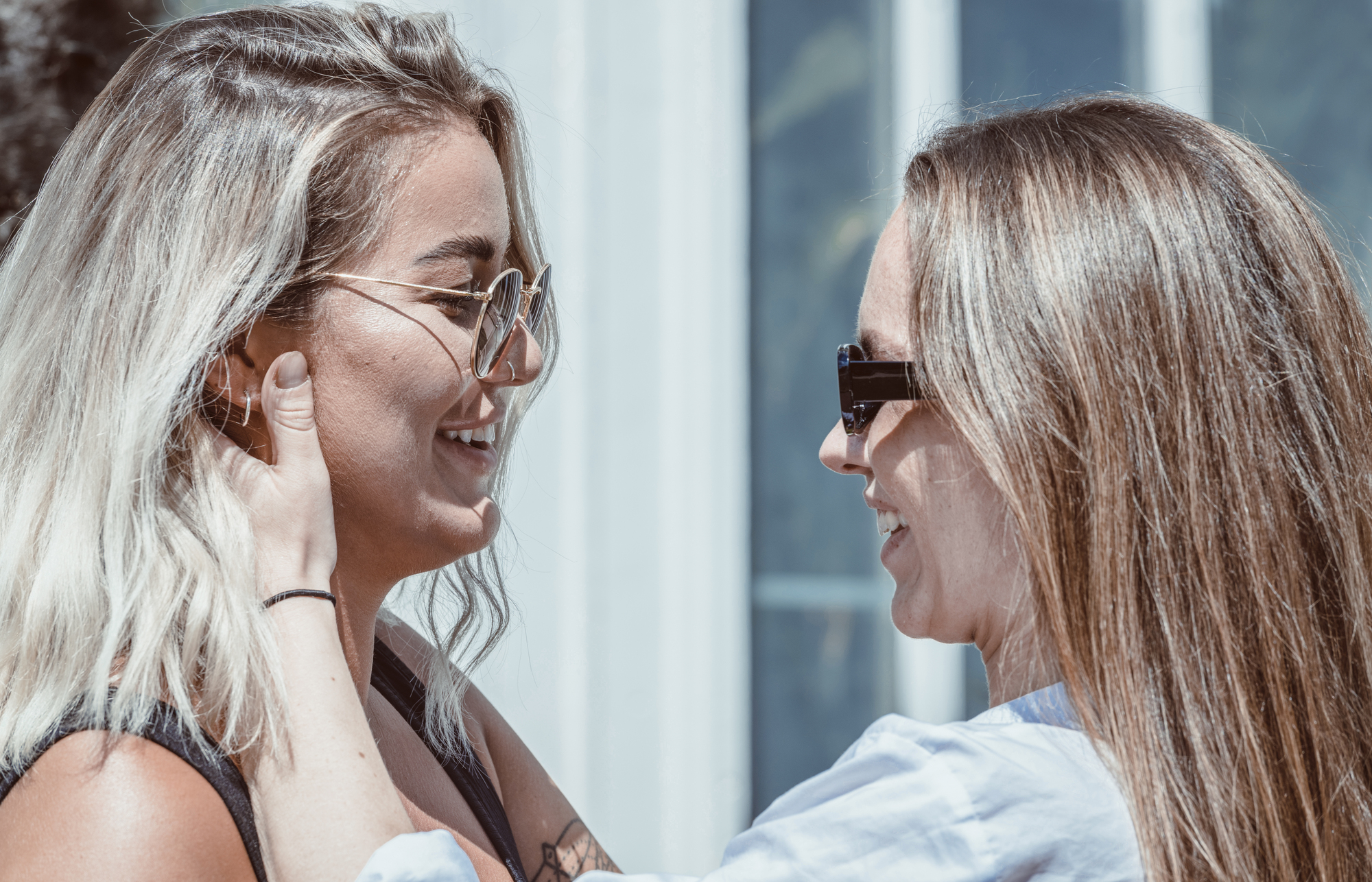 Two women stand facing each other, both wearing sunglasses. One woman has light blonde hair and the other has long, light brown hair. They appear to be in a happy, affectionate interaction. One woman gently holds the other's face, and they are smiling.