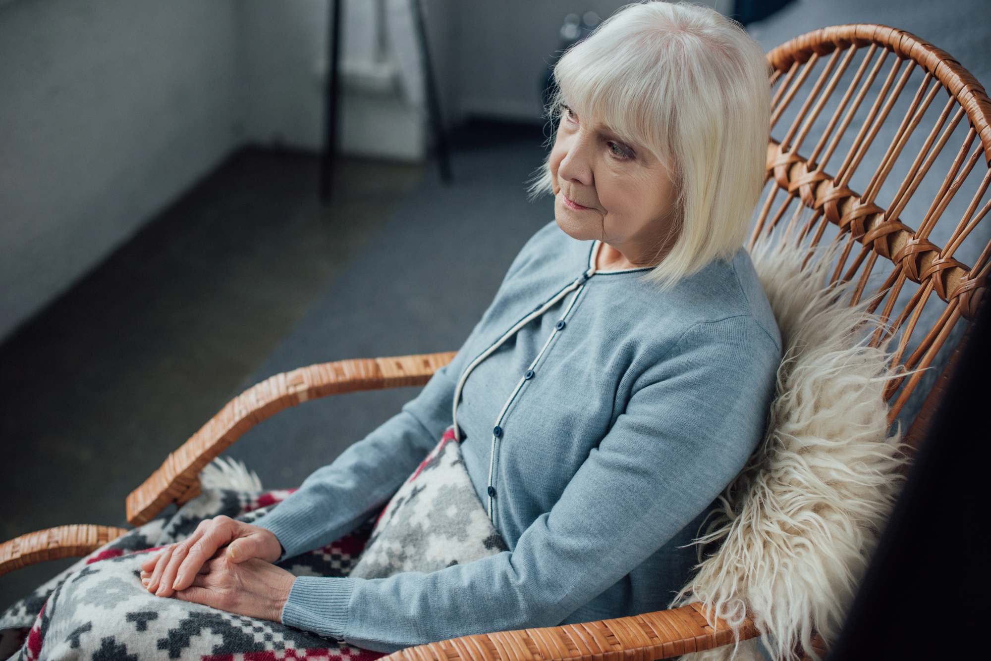 An elderly woman with white hair sits pensively in a wicker chair. She is wearing a light blue cardigan and has a blanket with a geometric pattern draped over her lap. A fluffy cushion is on the back of her chair and the room appears cozy and serene.