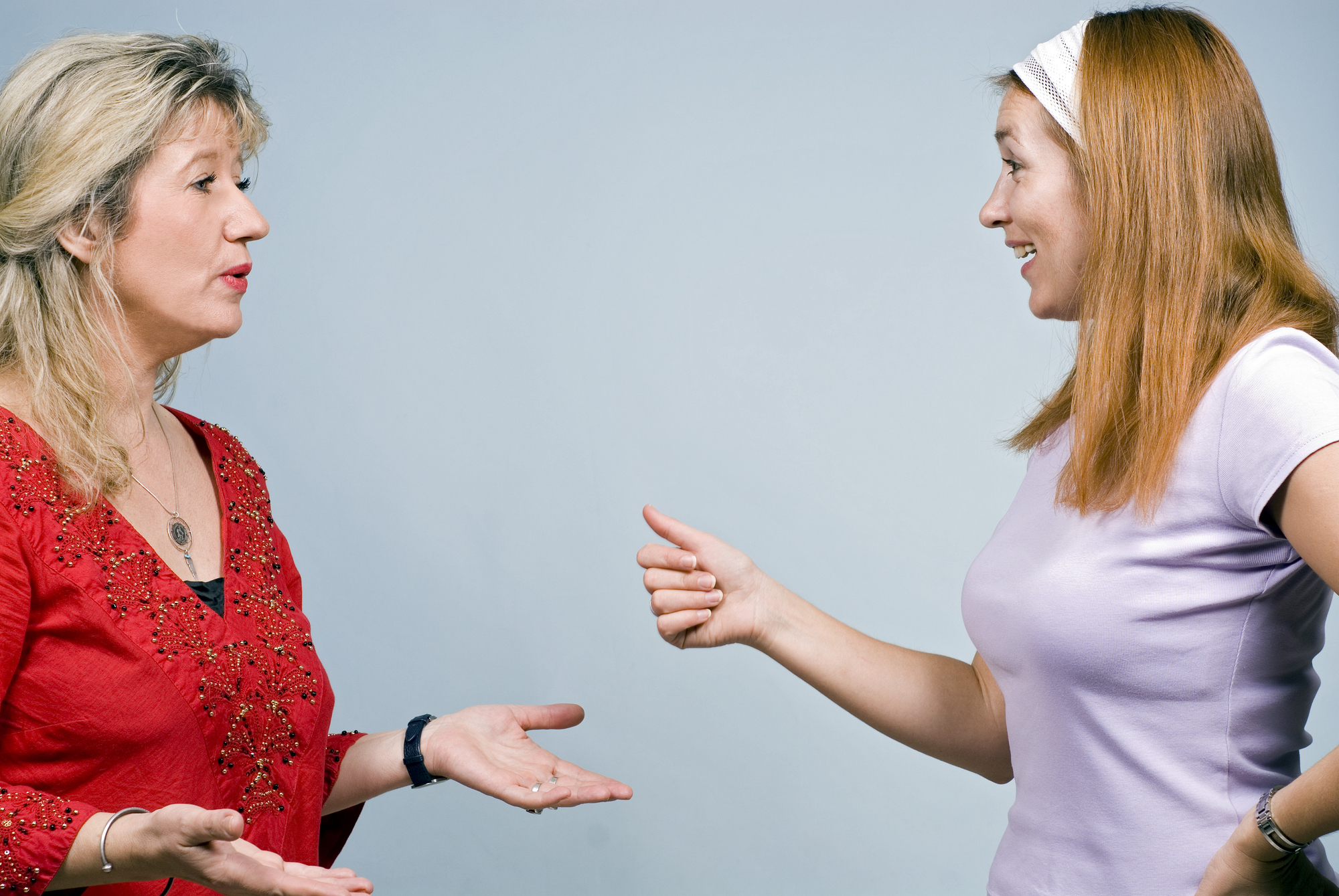 Two women engage in a conversation against a plain blue background. One woman, wearing a red embroidered top, gestures with open hands. The other woman, in a white headband and a light purple shirt, responds with a pointing gesture and a smile.