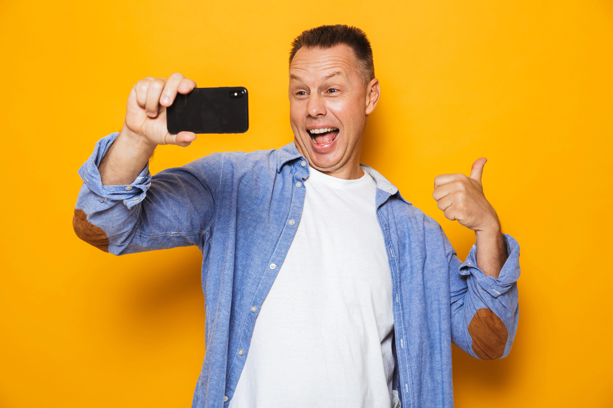 A cheerful man in a blue shirt and white T-shirt is taking a selfie with his smartphone against a bright yellow background. He is smiling widely and giving a thumbs-up with his other hand.