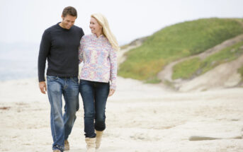 A man and woman are walking arm in arm on a sandy beach. Both are dressed in casual sweaters and jeans. The woman is wearing boots, and the man is in shoes. The background shows greenery and a cloudy sky. They appear to be enjoying a pleasant, relaxed stroll.