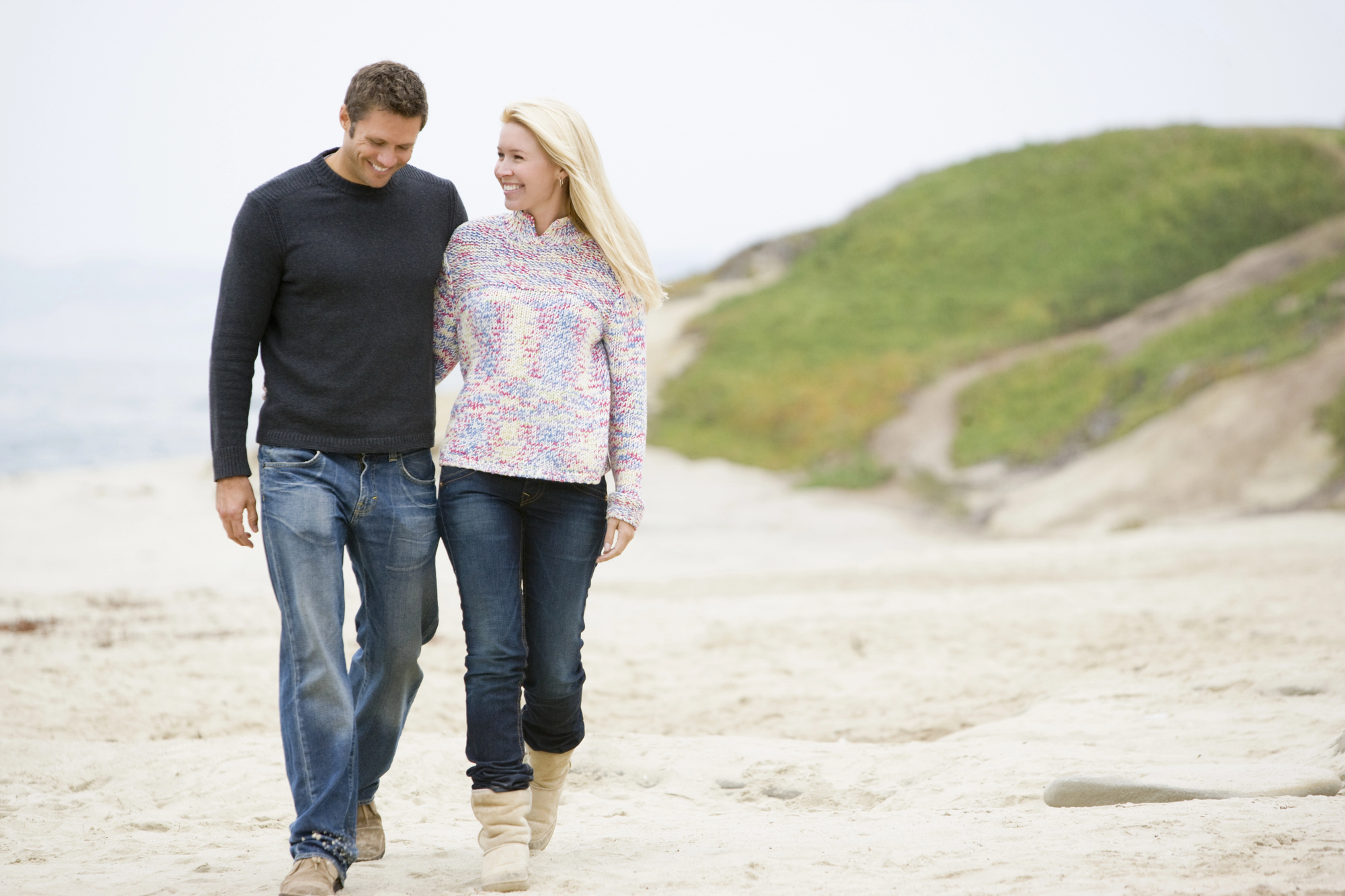 A man and woman are walking arm in arm on a sandy beach. Both are dressed in casual sweaters and jeans. The woman is wearing boots, and the man is in shoes. The background shows greenery and a cloudy sky. They appear to be enjoying a pleasant, relaxed stroll.