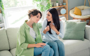 Two women are sitting on a light-colored sofa in a cozy living room. One has short hair and wears a green shirt, while the other has long hair and wears a light blue shirt. They seem to be having an emotional conversation, with one woman placing her hand on the other's arm.