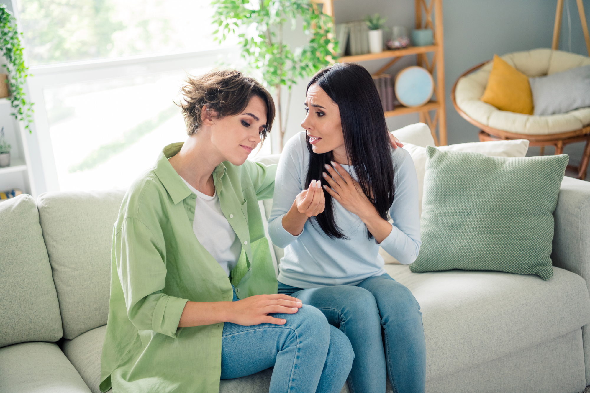 Two women are sitting on a light-colored sofa in a cozy living room. One has short hair and wears a green shirt, while the other has long hair and wears a light blue shirt. They seem to be having an emotional conversation, with one woman placing her hand on the other's arm.