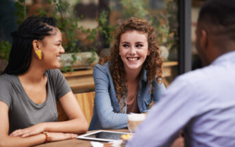 Three people are sitting at an outdoor café, engaged in a lively conversation. One person is facing away from the camera, while the other two, a woman with curly hair and a woman with braids, are smiling at each other. There are drinks and a tablet on the table.