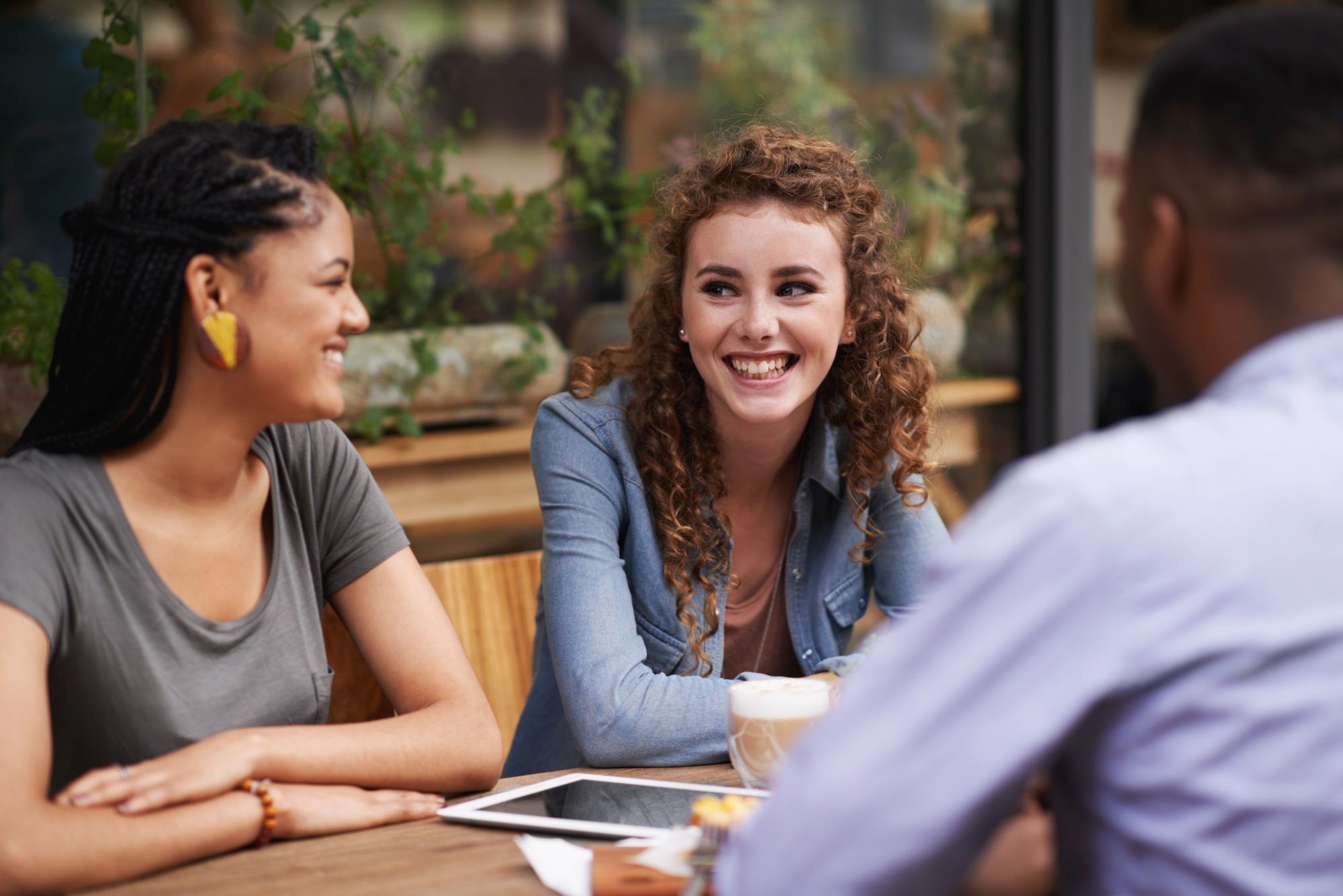 Three people are sitting at an outdoor café, engaged in a lively conversation. One person is facing away from the camera, while the other two, a woman with curly hair and a woman with braids, are smiling at each other. There are drinks and a tablet on the table.