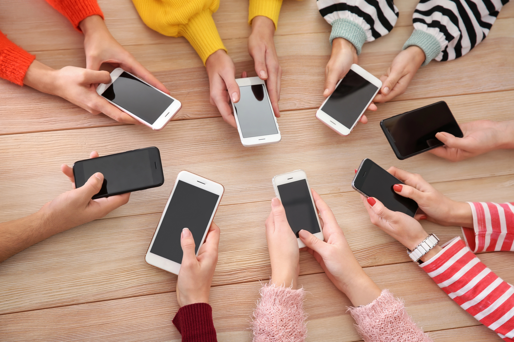 A group of people sit around a table, each holding a smartphone in their hands. The phones have various case designs, and the individuals' clothing sleeves create a colorful array. The table is wooden and the devices are all turned on with their screens lit.