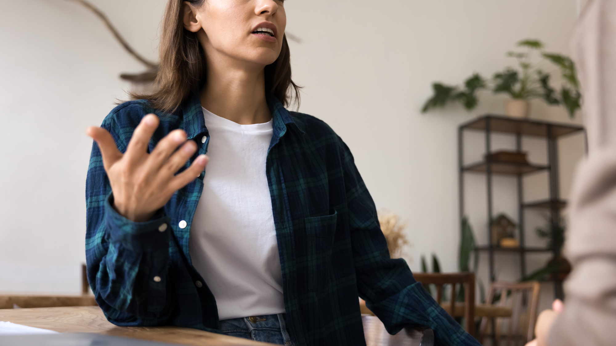 A person with long hair wearing a white T-shirt and blue plaid shirt gestures with their right hand while speaking. They are indoors, and behind them are shelves with potted plants and a table. The image captures a moment of conversation.