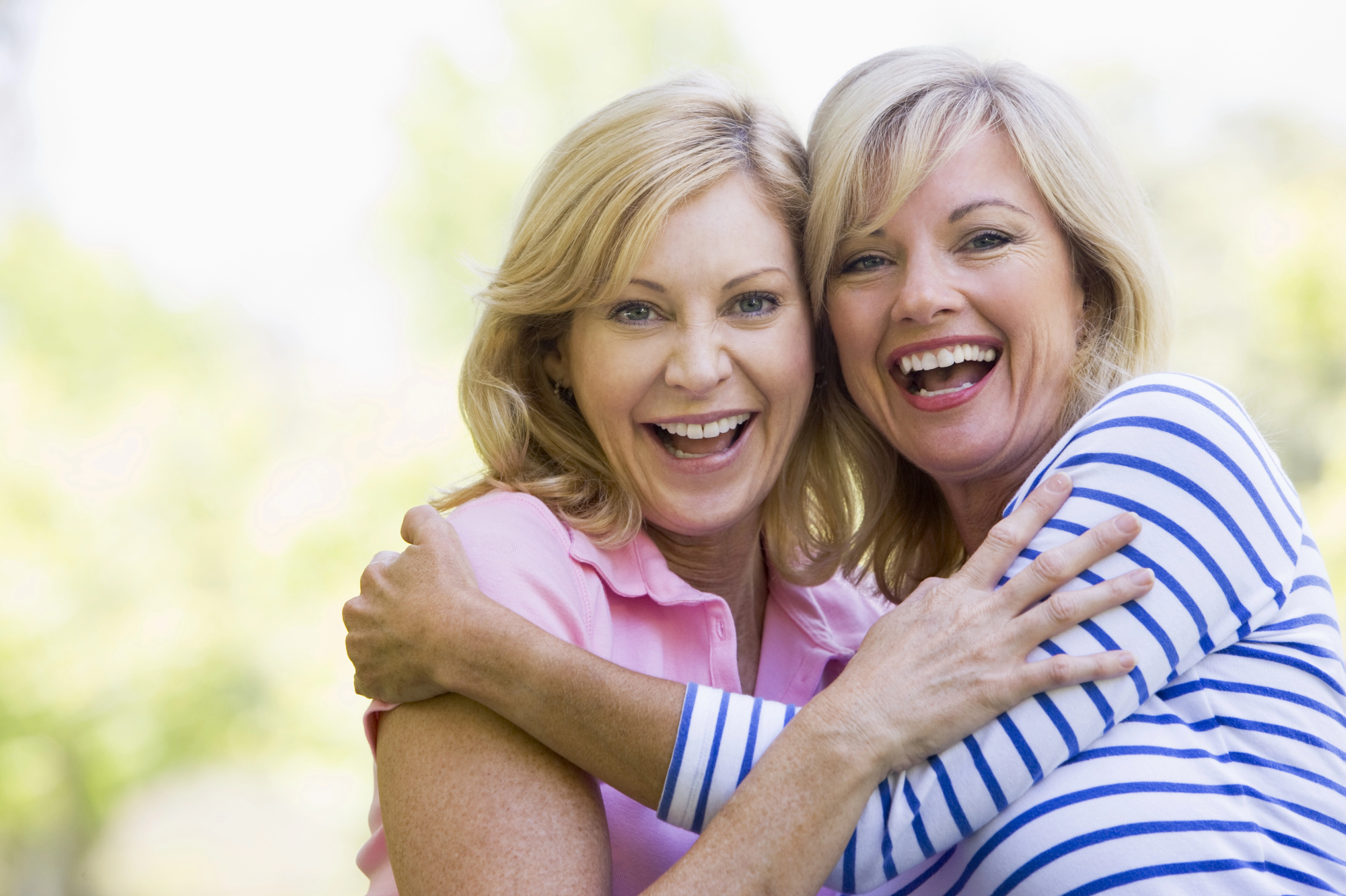 Two women are standing outdoors, smiling and hugging each other. The woman on the left is wearing a pink polo shirt, and the woman on the right is in a white and blue striped top. They appear cheerful and happy. The background is blurred and green, suggesting a park or natural setting.