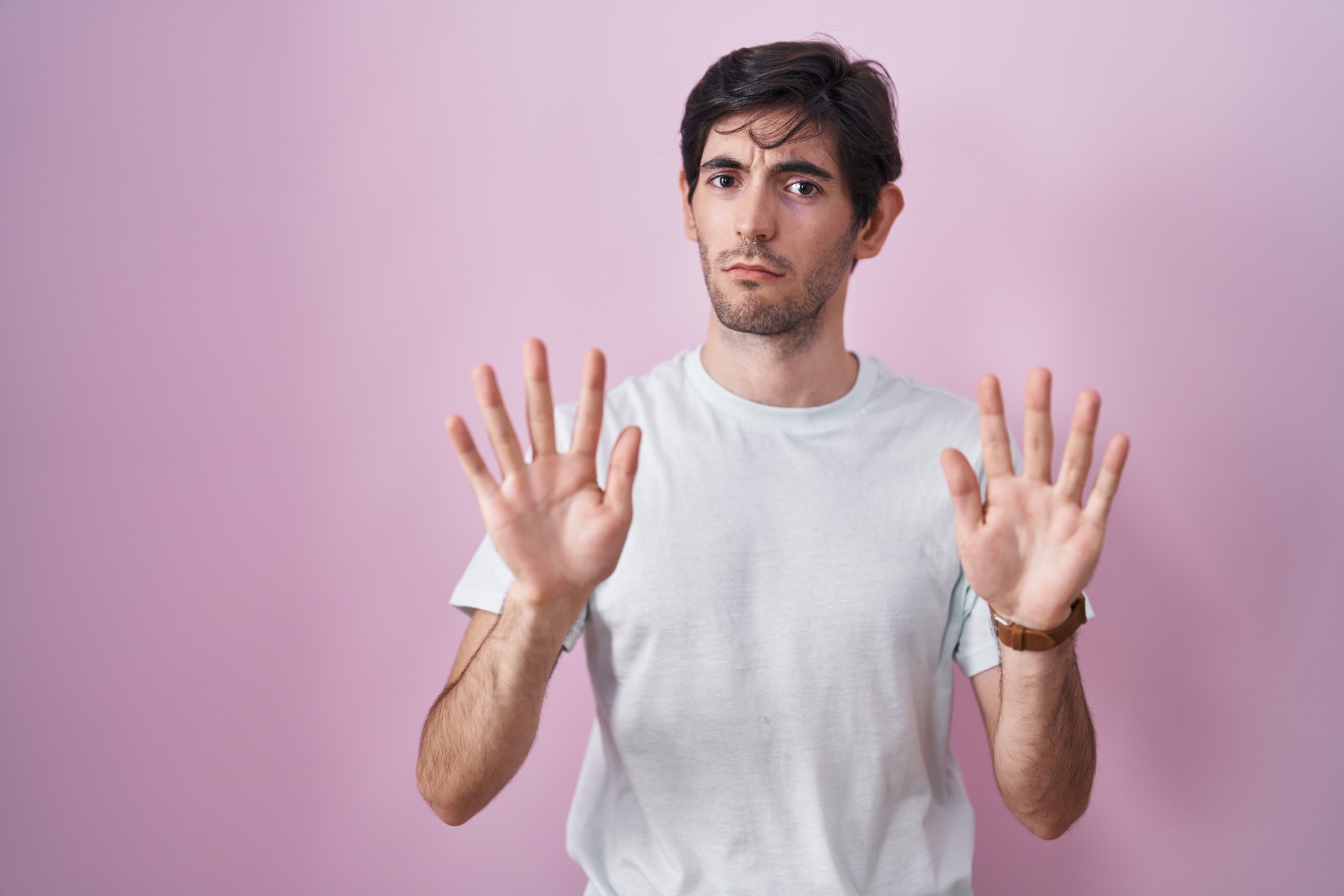 A man with dark hair and a beard is standing against a pink background. He is wearing a white t-shirt and has a serious expression on his face. Both of his hands are raised in front of him with palms facing outward, as if gesturing to stop or expressing caution.