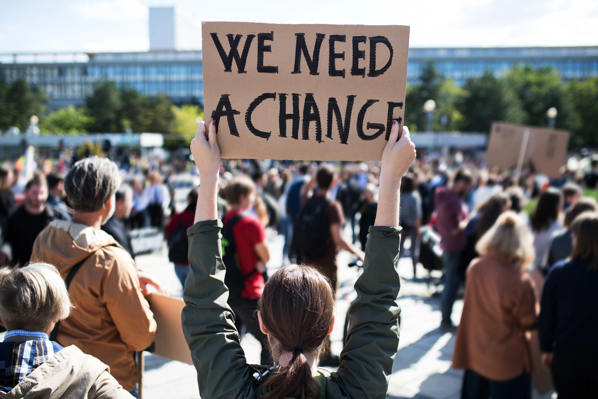 A person is holding up a cardboard sign that reads "WE NEED A CHANGE" at a protest. The person stands in the foreground with their back to the camera, and a large crowd of protesters with various signs is gathered in the background.