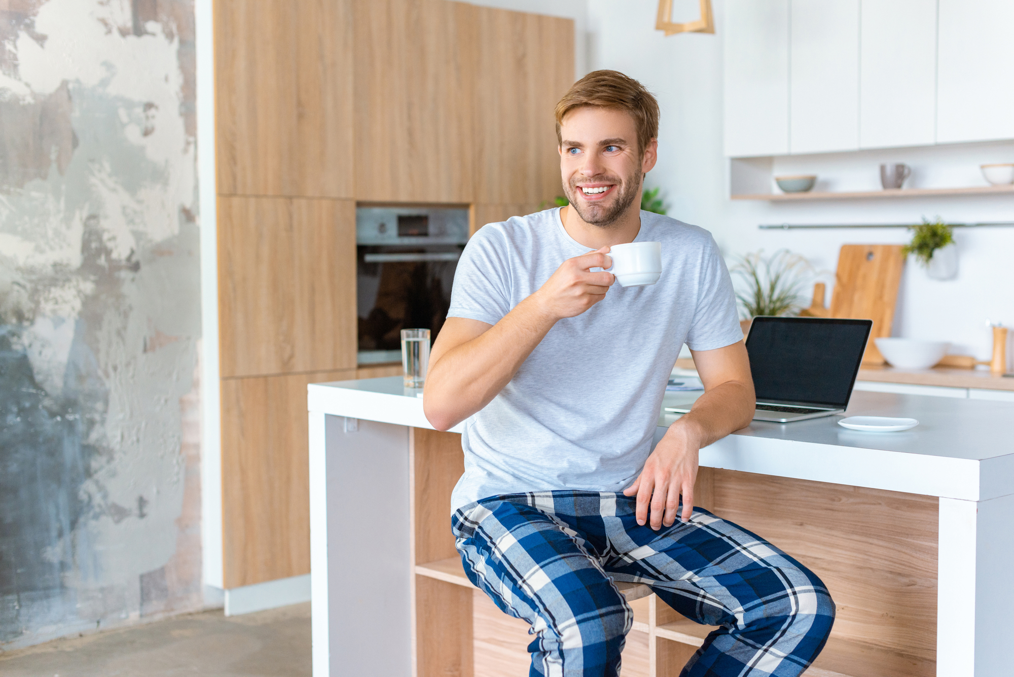 A man with light brown hair and a beard, dressed in a gray t-shirt and blue plaid pajama pants, sits on a kitchen counter enjoying a cup of coffee. Behind him is a modern kitchen with white cabinets and wooden accents, along with a laptop on the counter.