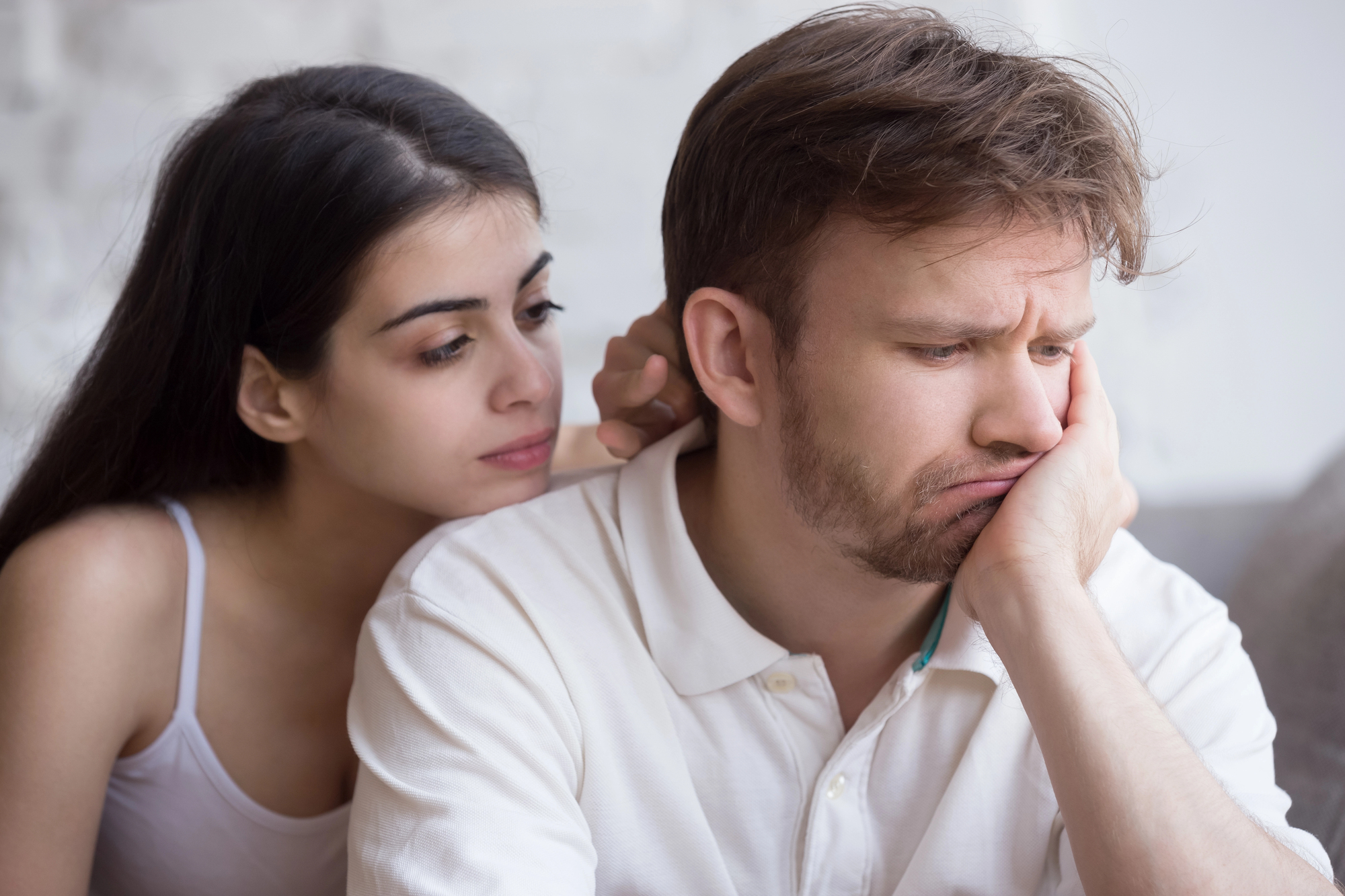 A woman with long dark hair consoles a man with short brown hair and a beard. The man, dressed in a white shirt, appears sad and thoughtful, resting his face in his hand. The woman, wearing a white tank top, looks at him sympathetically.