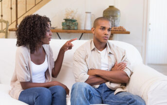 A woman is sitting on a couch, pointing her finger and talking to a man next to her who has his arms crossed and an unhappy expression. Both are casually dressed in a white T-shirt and jeans. The background shows a decorated living room.