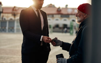 In a city square, a man in a suit and tie hands money to an older man wearing a red beanie and gloves, who holds a cup. The older man appears to be in need, while the suited man stands in a compassionate gesture. Buildings and sunlight are visible in the background.