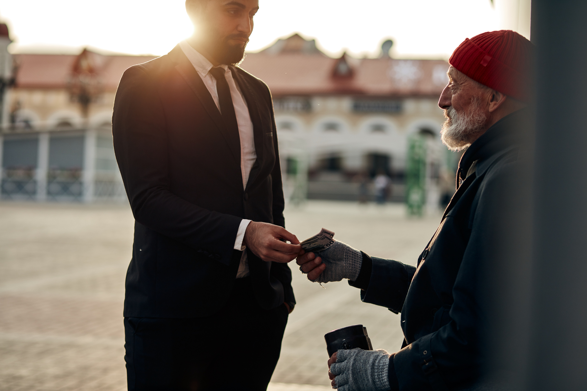 In a city square, a man in a suit and tie hands money to an older man wearing a red beanie and gloves, who holds a cup. The older man appears to be in need, while the suited man stands in a compassionate gesture. Buildings and sunlight are visible in the background.