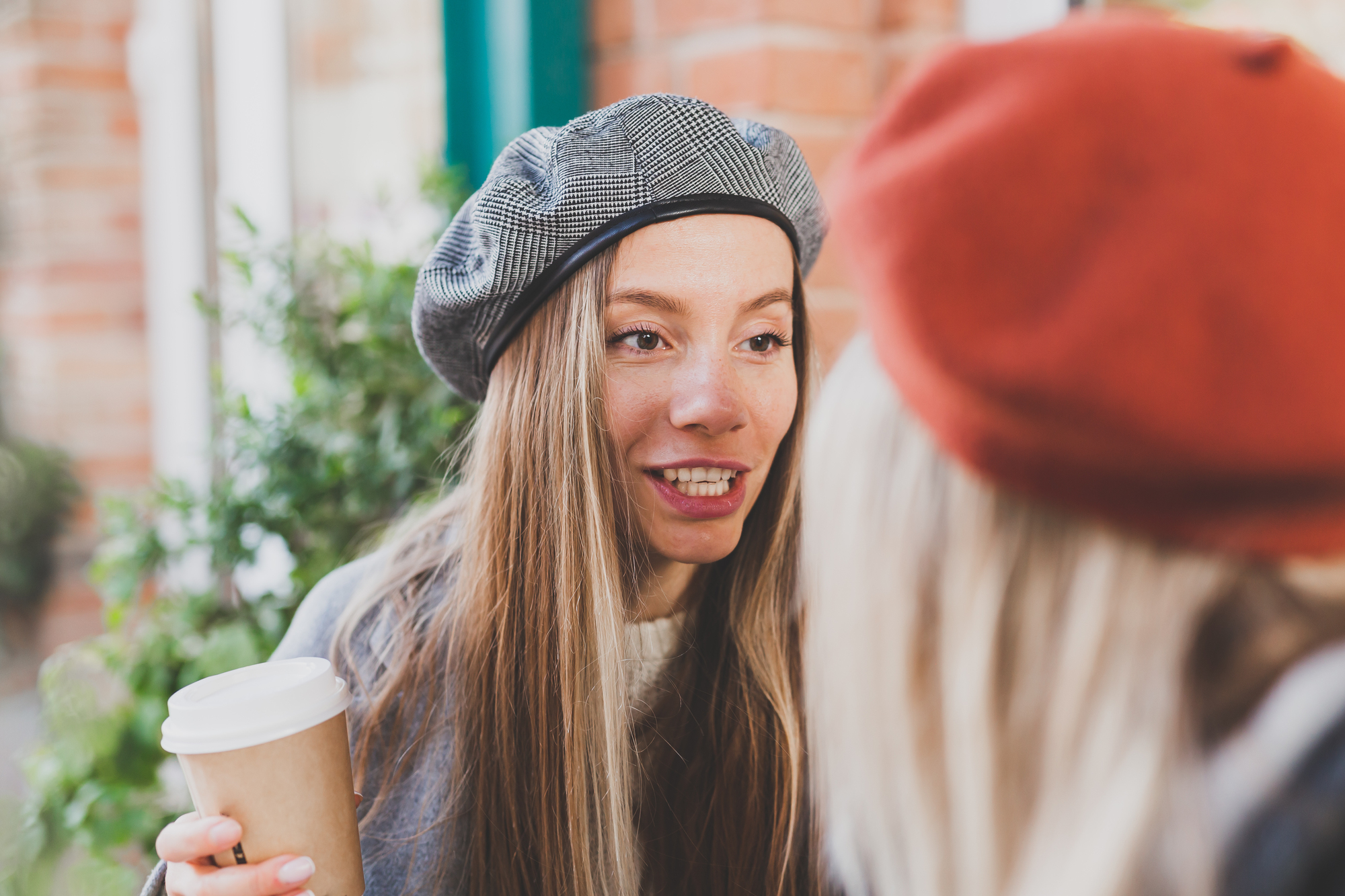 A woman wearing a gray beret and holding a paper coffee cup is speaking to another person, whose face is not visible, but their red beret is in focus. They are outside, with a blurred background of greenery and a brick building.