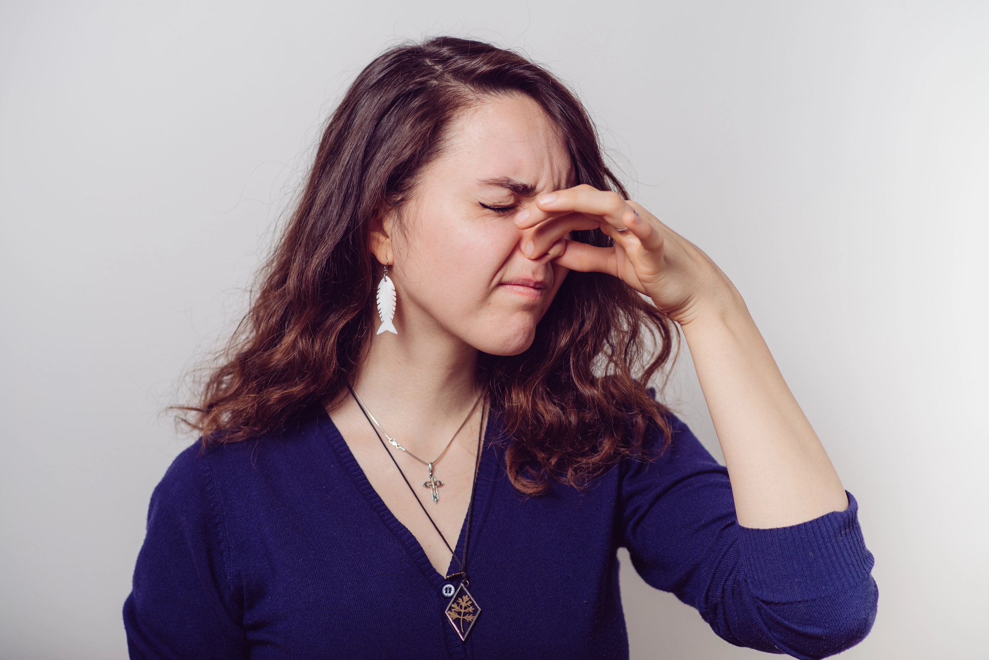 A woman in a blue sweater and wearing a necklace and earrings is standing against a plain background, pinching her nose with a displeased expression, suggesting she smells something unpleasant. Her eyes are closed, and her head is tilted slightly downward.