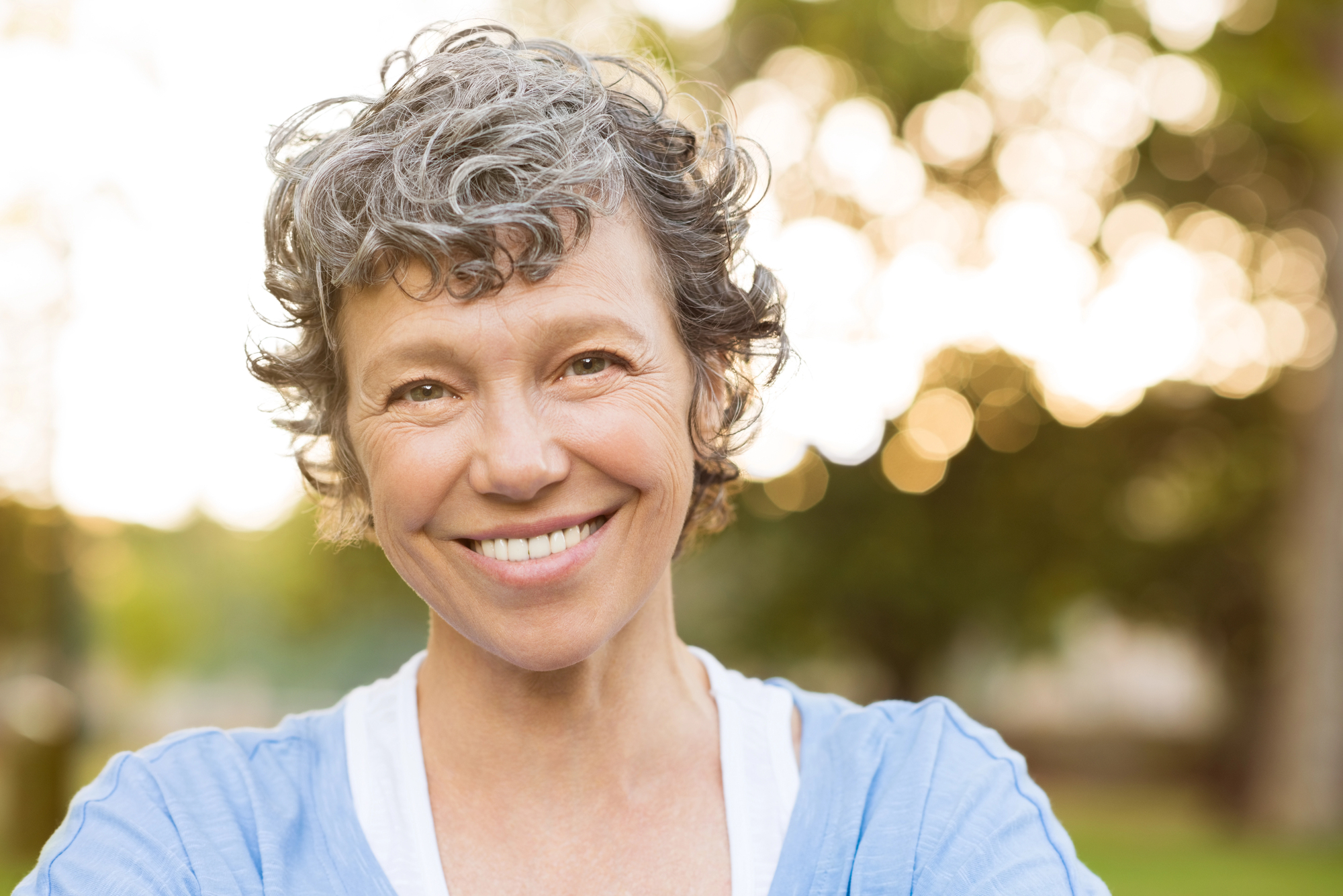 Smiling elderly woman with curly gray hair, wearing a blue shirt, standing outdoors with blurred greenery and sunlight in the background.