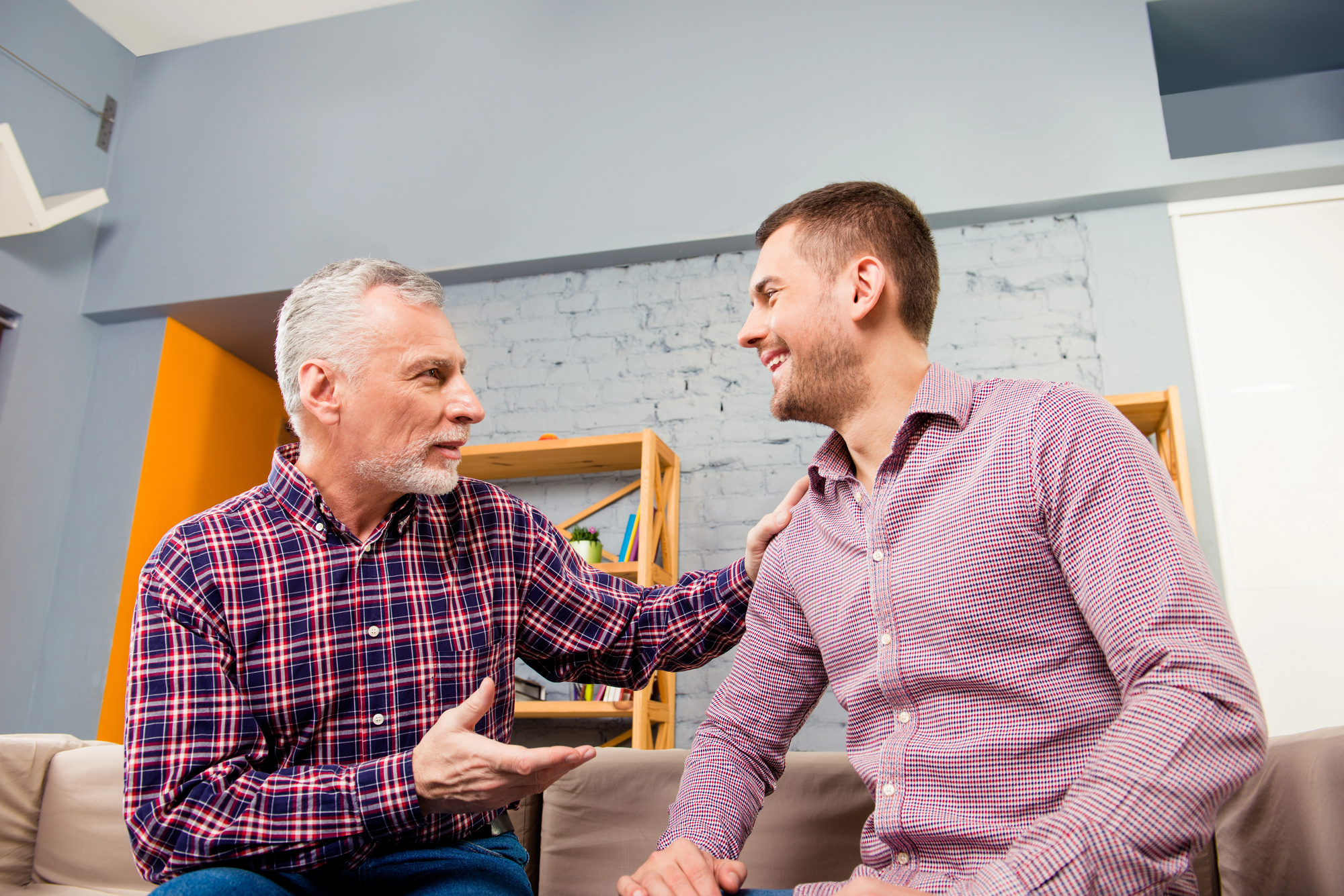 Two men are having a conversation in a living room. The older man on the left, with gray hair and a plaid shirt, is gesturing with one hand while his other hand rests on the younger man's shoulder. The younger man, with short hair and a striped shirt, is smiling.