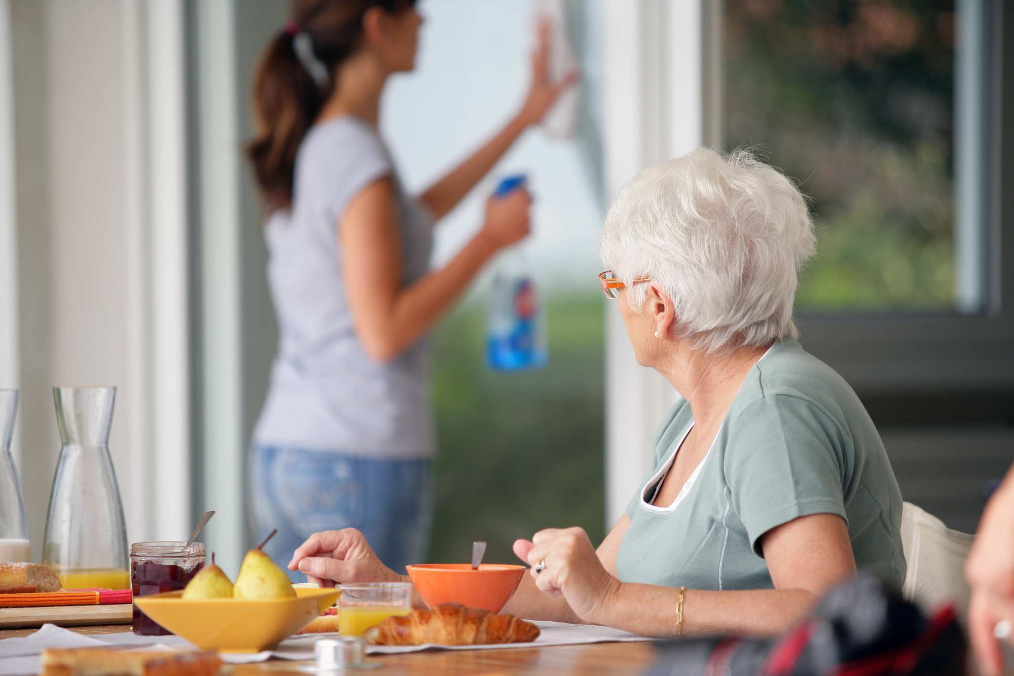 An elderly woman with short white hair sits at a table set with breakfast items, including a bowl, croissant, and fruit. She looks at a younger woman in the background who is cleaning a window with a spray bottle. The scene appears to be indoors.