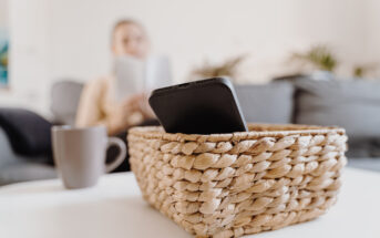 A smartphone placed in a woven basket on a white table, with a blurred background showing a person reading a book on a gray couch and a gray mug nearby.