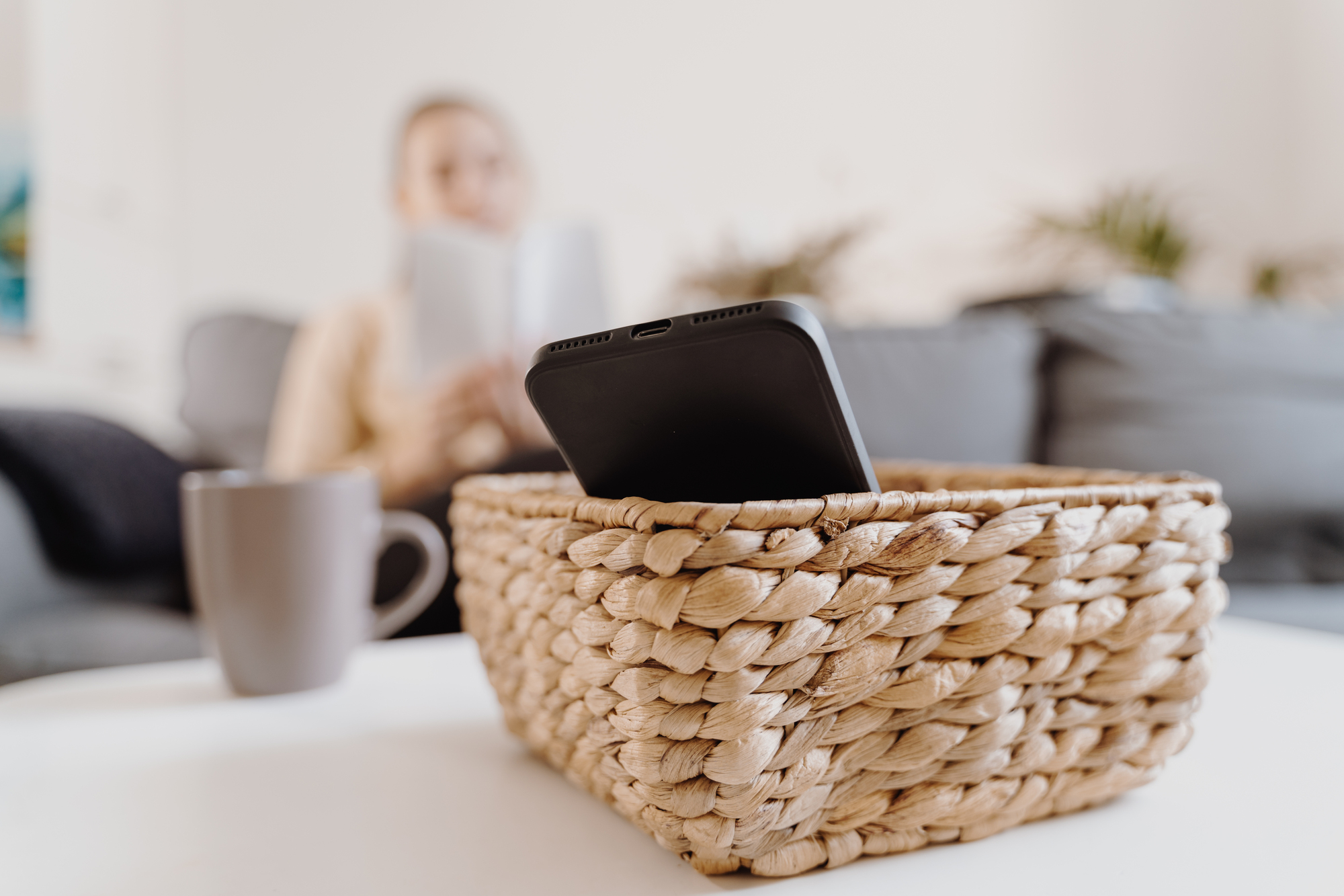 A smartphone placed in a woven basket on a white table, with a blurred background showing a person reading a book on a gray couch and a gray mug nearby.