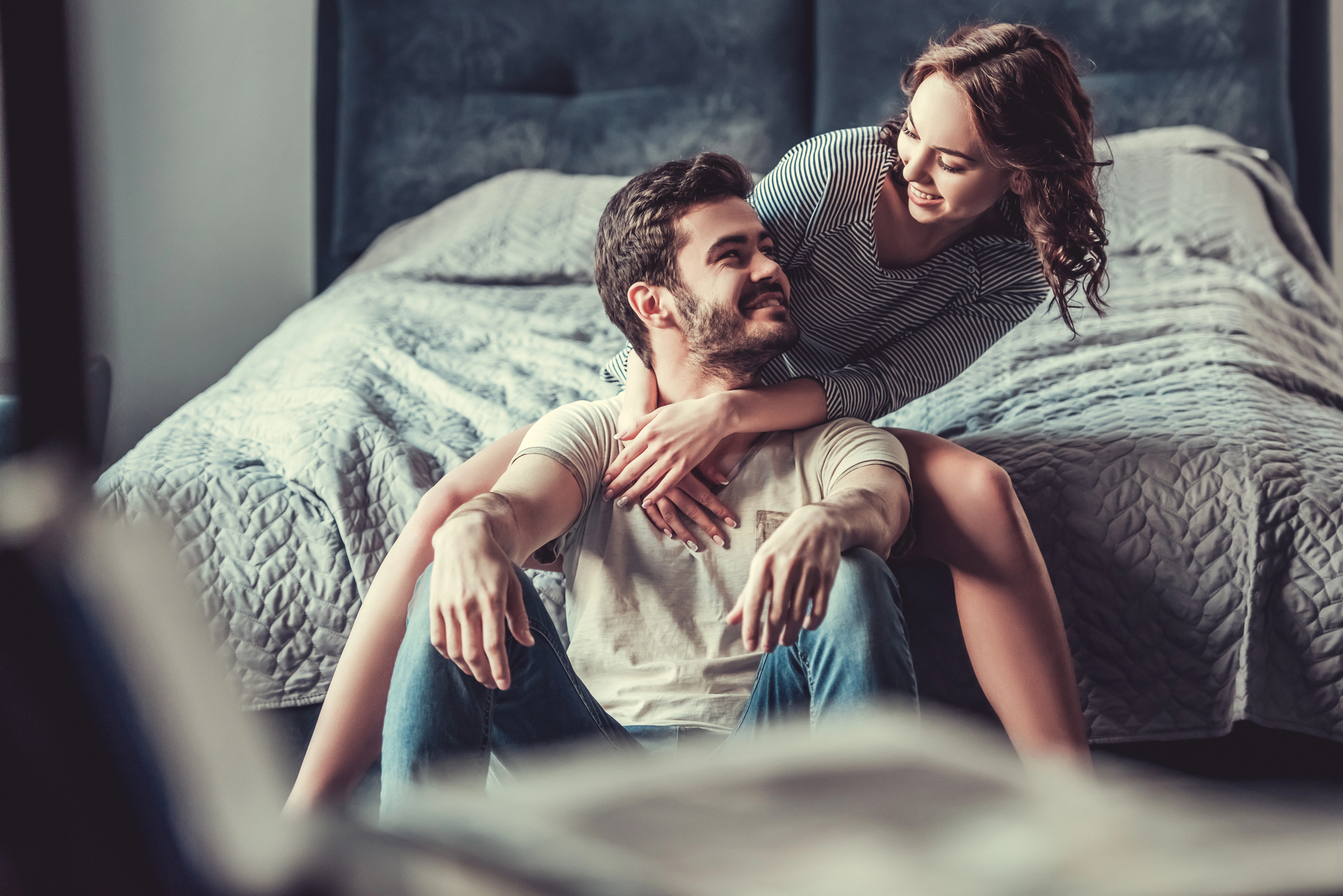 A couple sits on the floor in front of a bed. The man is seated with his legs stretched out, and the woman kneels behind him, hugging him around the neck and smiling. The room has a cozy, intimate atmosphere with soft lighting.