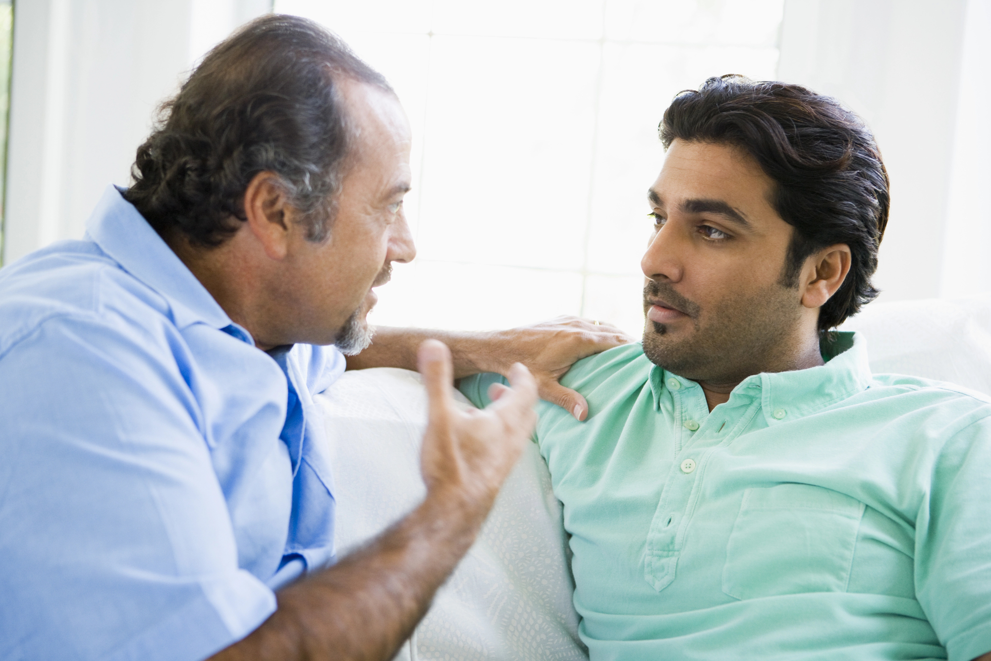 A middle-aged man with gray hair wearing a light blue shirt is sitting close to a younger man with dark hair and a green shirt. They appear to be having a serious conversation, with the older man gesturing with his hand while the younger man listens intently.