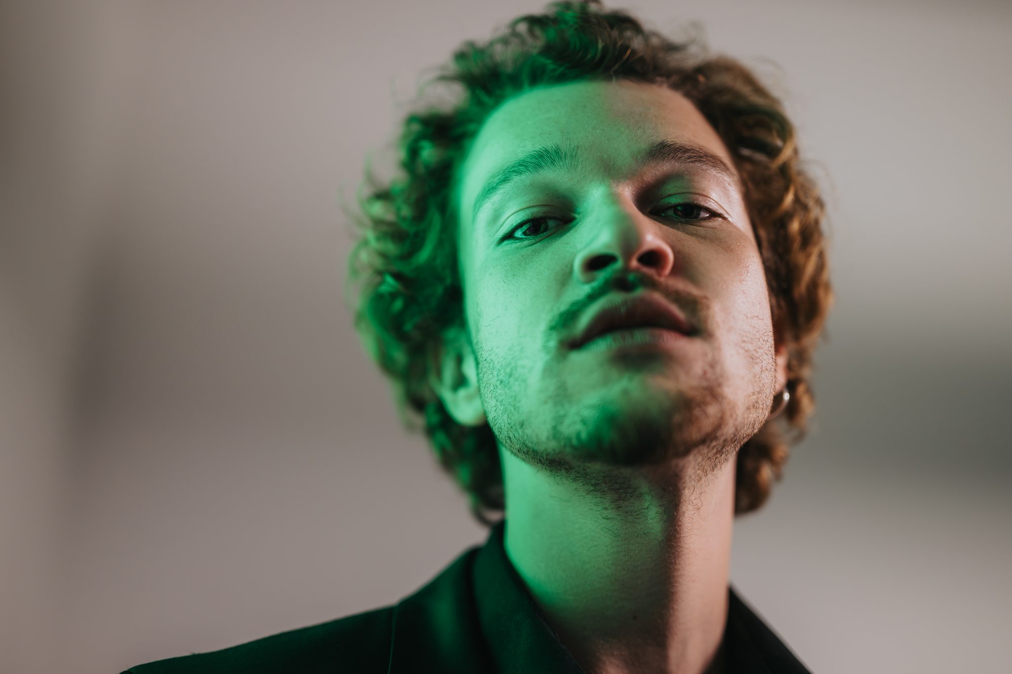 A person with curly hair and a beard looks downward into the camera under green lighting, creating soft shadows on their face. They wear a dark jacket and have a serious expression, with a blurred background enhancing the focus on their face.