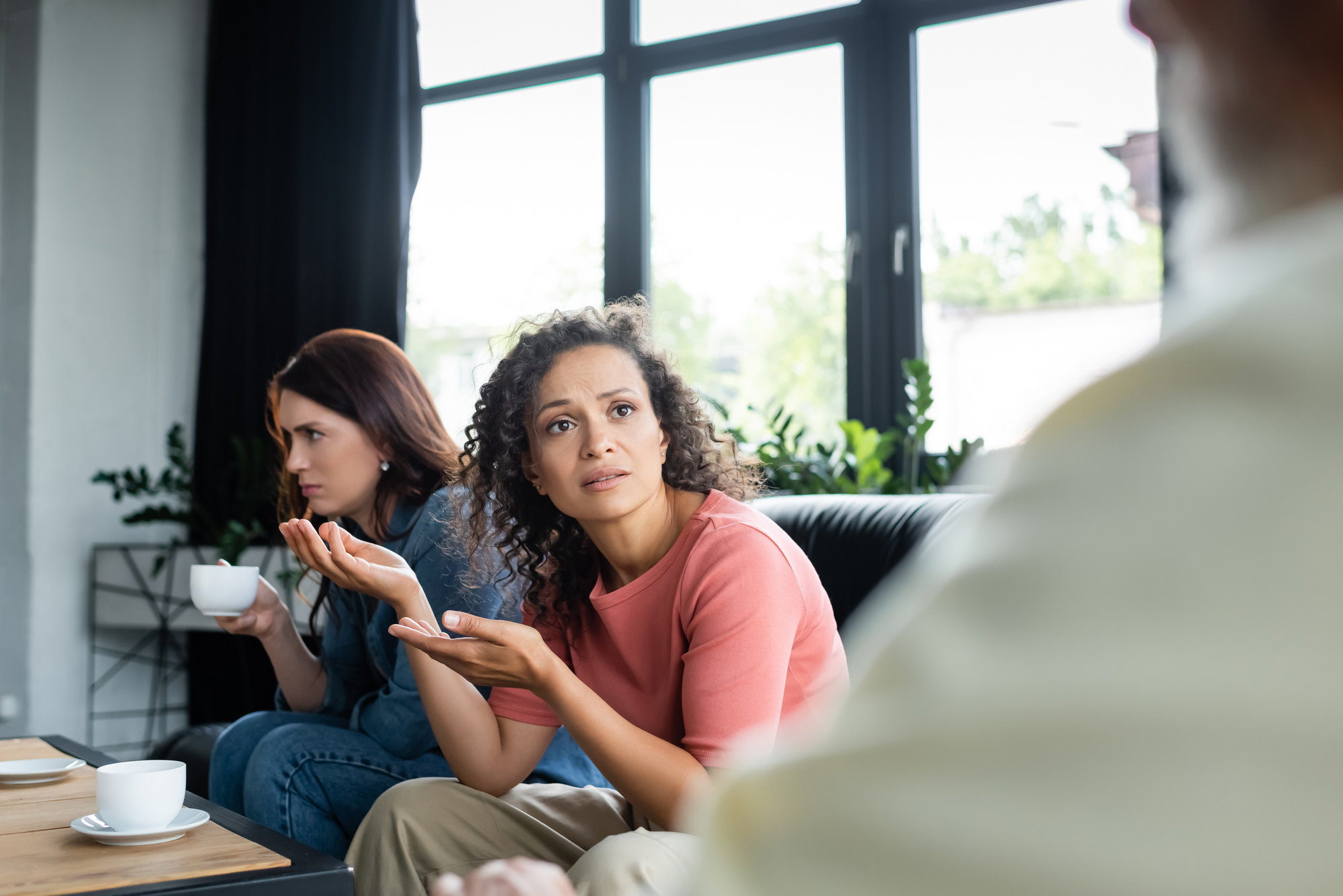 Two women sit on a sofa in a living room. The woman on the right, with curly hair and wearing a pink shirt, gestures with her hands while looking concerned. The woman on the left, with straight hair and wearing a denim jacket, holds a cup with both hands and looks away.