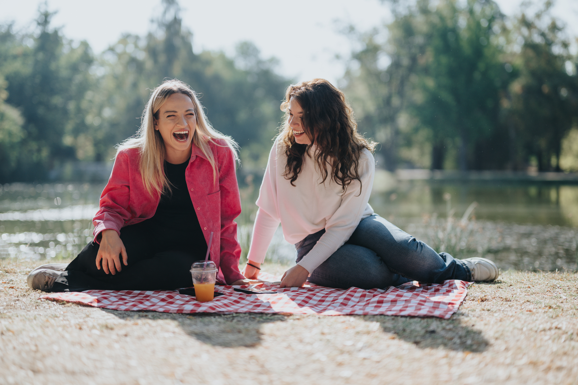 Two women sitting on a red checkered picnic blanket by a lake, laughing joyfully under the bright sunshine. One has blonde hair and wears a pink jacket, while the other has brown hair and wears a white sweater. An orange drink in a plastic cup sits on the blanket.