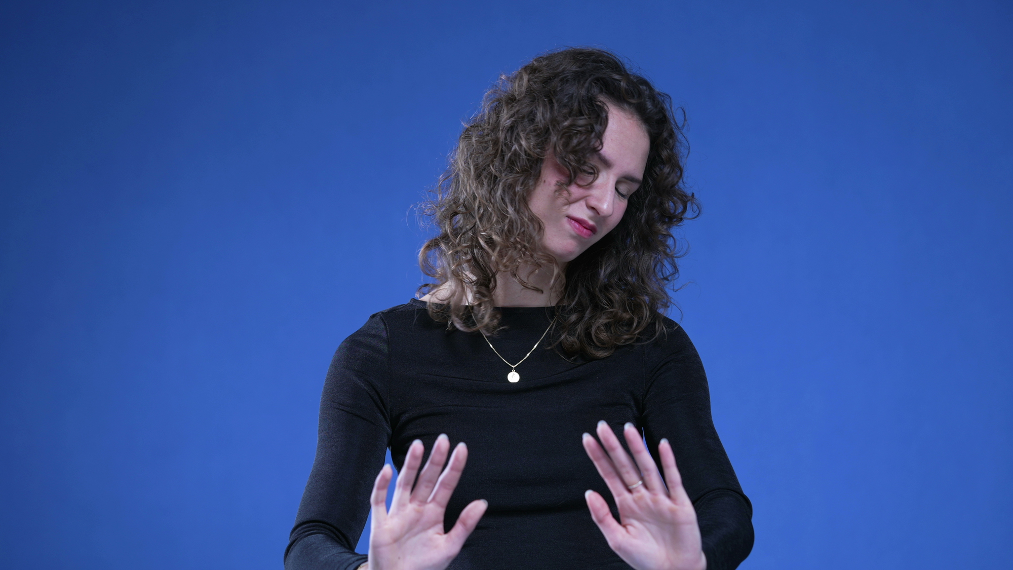 A woman with curly hair, wearing a black long-sleeve top and a pendant necklace, stands against a blue background. She has her eyes closed, with her hands raised and palms facing outward, giving the impression of stopping or rejecting something.