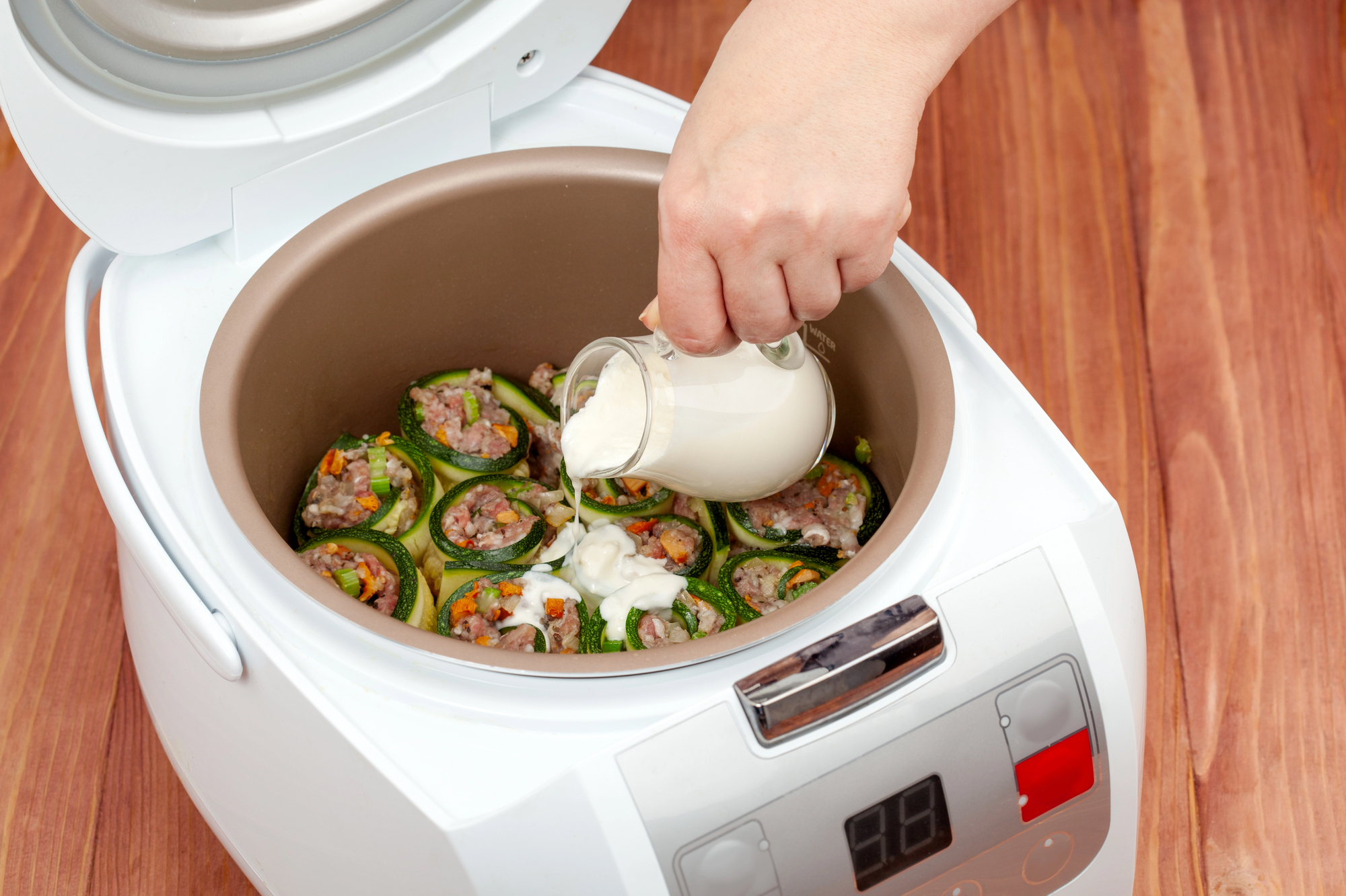 A slow cooker with stuffed zucchini rolls being prepared. A hand is pouring creamy sauce from a small pitcher over the rolls. The slow cooker is on a wooden countertop, and its digital display shows it is preheating.