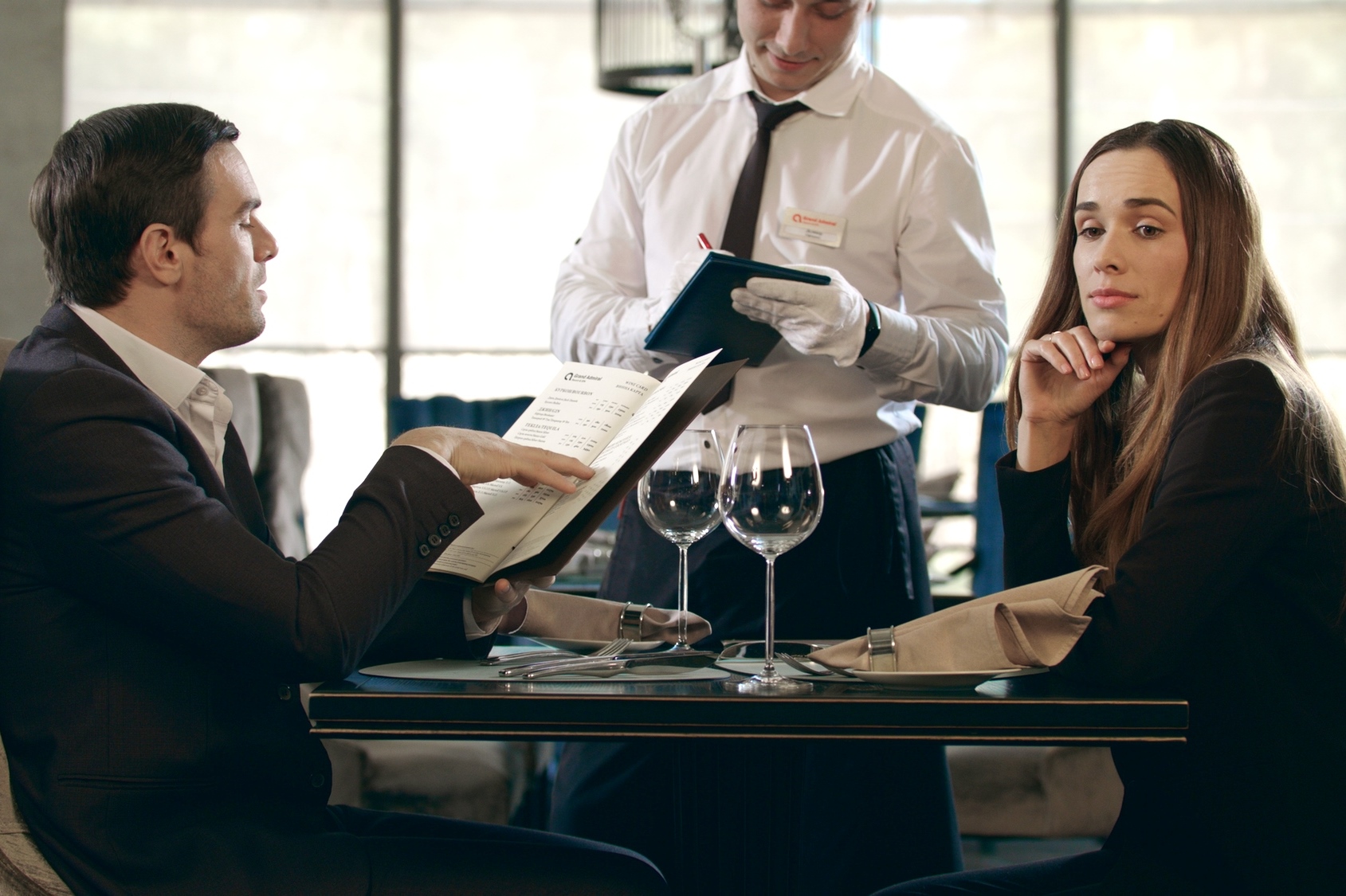 A man and a woman in business attire are sitting at a dining table in a restaurant, with wine glasses and menus in front of them. The man is looking at the menu, while the woman is looking at the camera with a thoughtful expression. A waiter is taking their order.