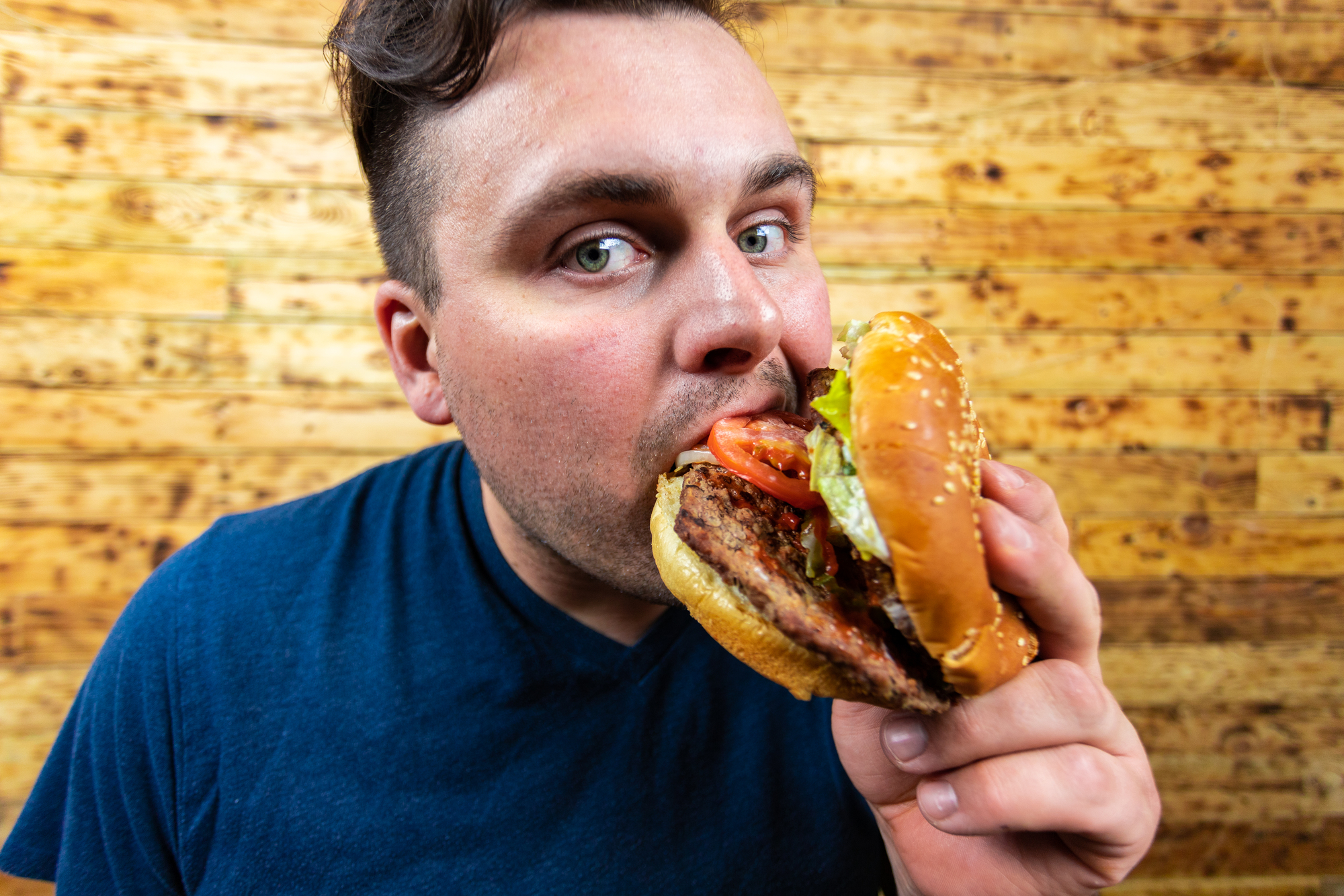 A person in a blue shirt is close to taking a bite from a large sandwich with a sesame seed bun, beef patty, tomato slices, and green lettuce. The background consists of wooden planks.