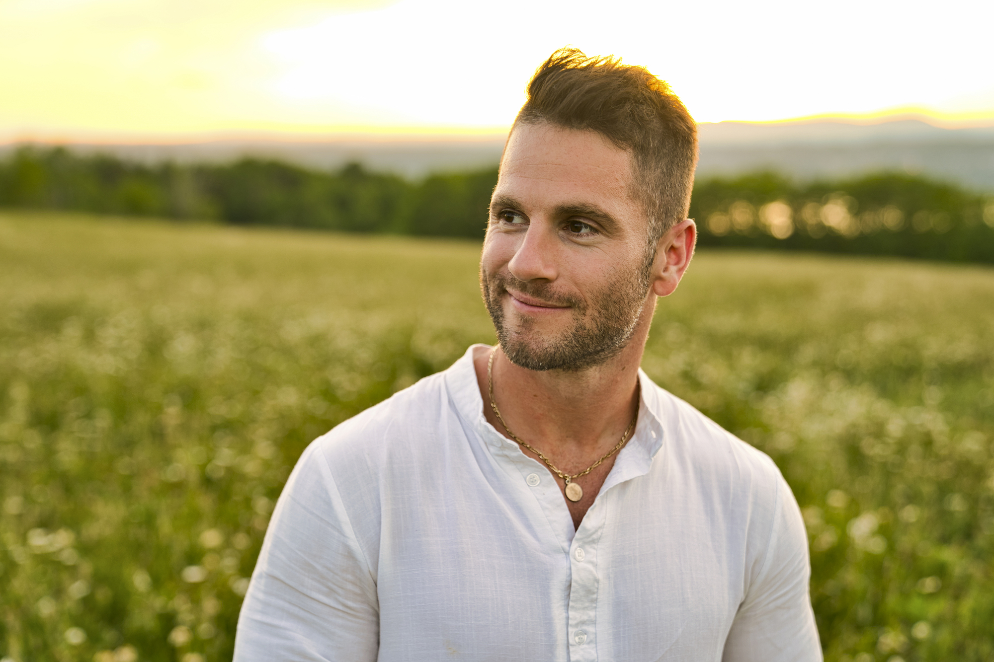 A man with short hair and a trimmed beard is standing in a field of tall grass, wearing a white shirt. The sun sets behind him, casting a warm glow over the landscape. He is smiling and looking slightly to the side. Trees and mountains are visible in the distance.