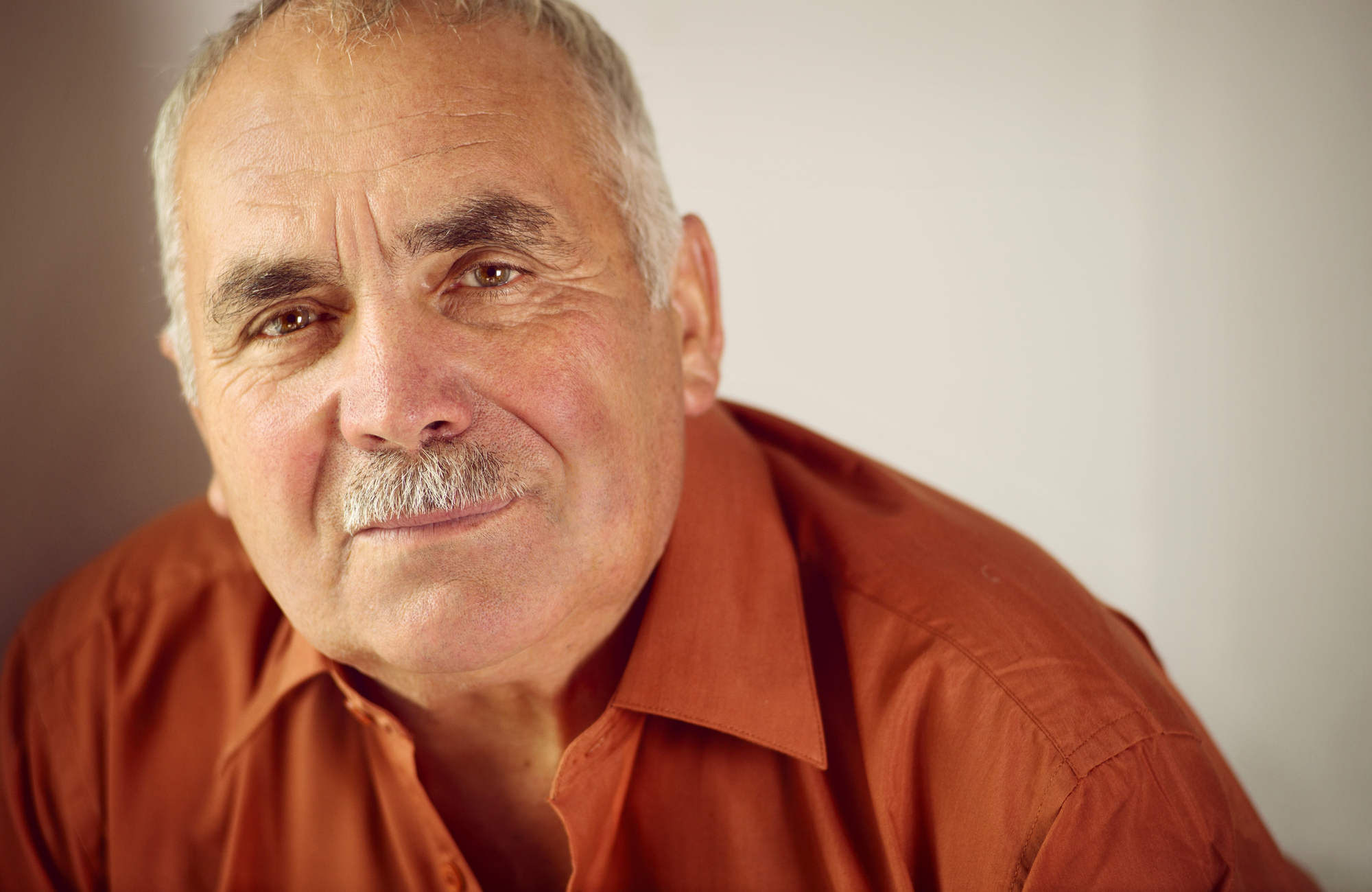 A close-up of an elderly man with short gray hair and a mustache, wearing an orange button-up shirt. The man has a gentle smile and is looking directly at the camera. The background is plain and softly lit.