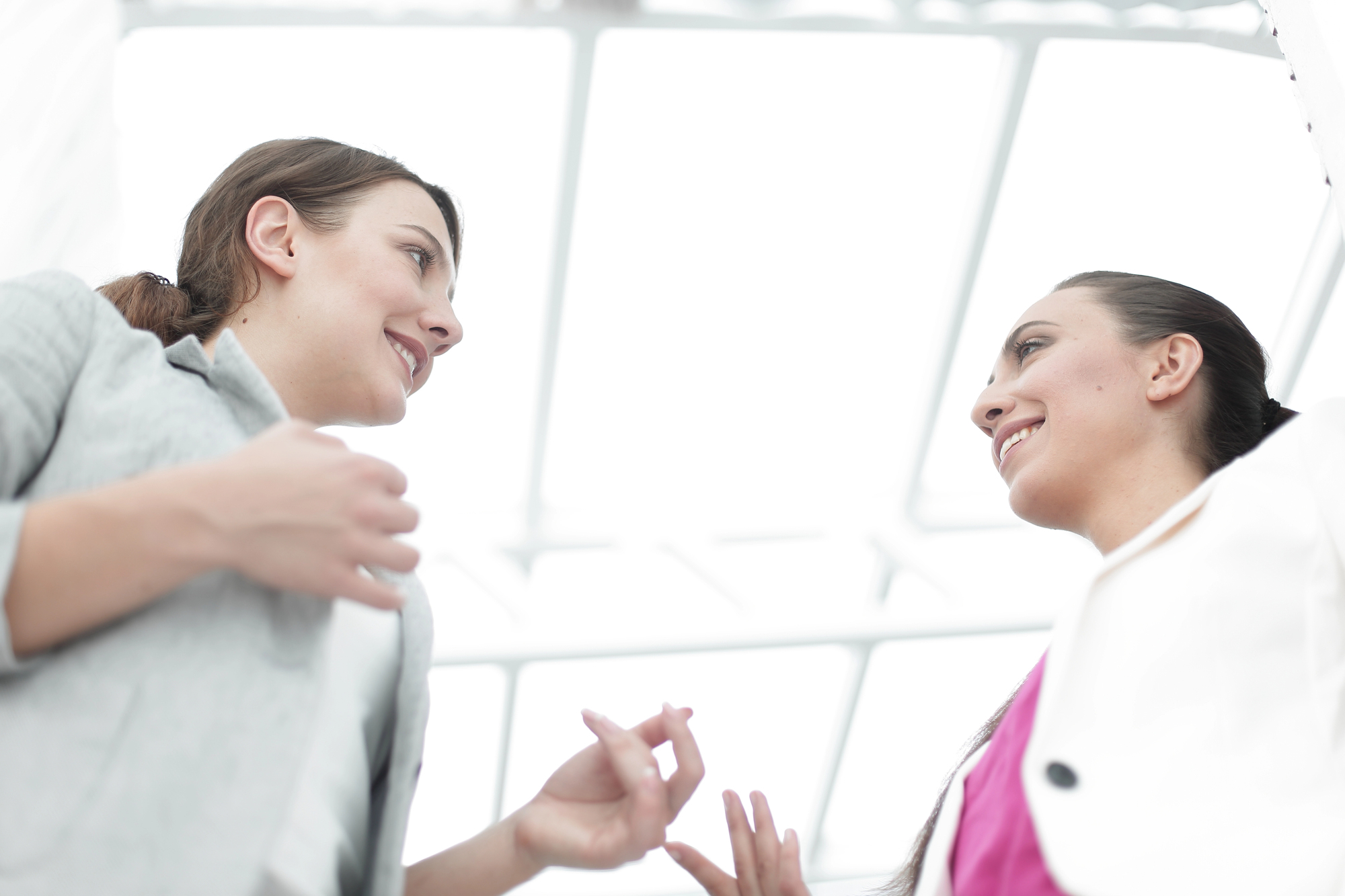 Two women engaged in a conversation in a bright, white space with a glass-paneled ceiling. They are smiling, one holding a piece of paper, both wearing professional attire. The image is taken from a low angle, emphasizing the open and airy environment.