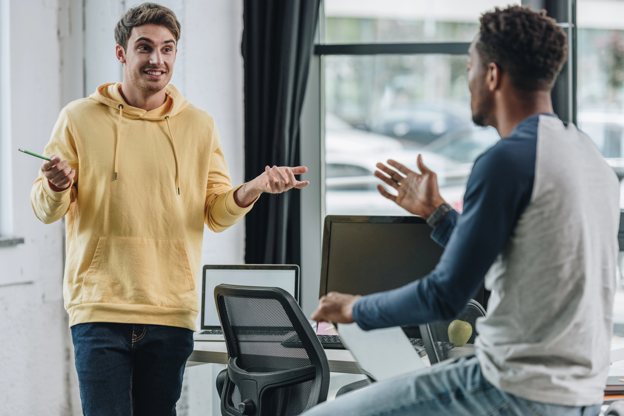 Two men engaged in a discussion in a modern office setting. One man in a yellow hoodie gestures while the other, in a gray and navy shirt, sits on a desk with a laptop in front of him. Both appear to be intently exchanging ideas or opinions.