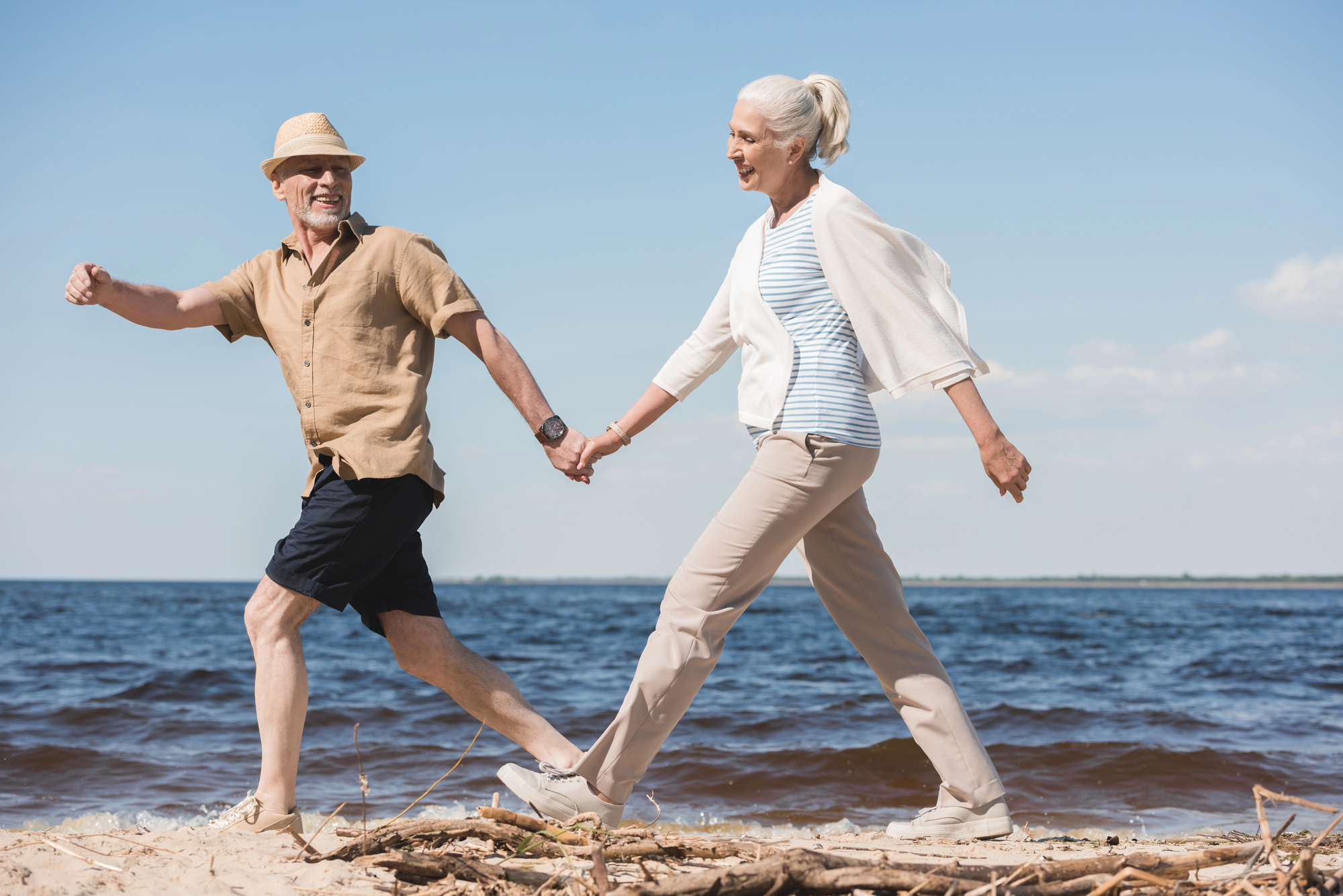 An elderly couple holding hands, joyfully walking along a beach. The man wears a hat, sunglasses, a beige shirt, and shorts. The woman has gray hair tied back, wearing a light cardigan over a striped shirt and beige pants. They smile, with the ocean and sky in the background.