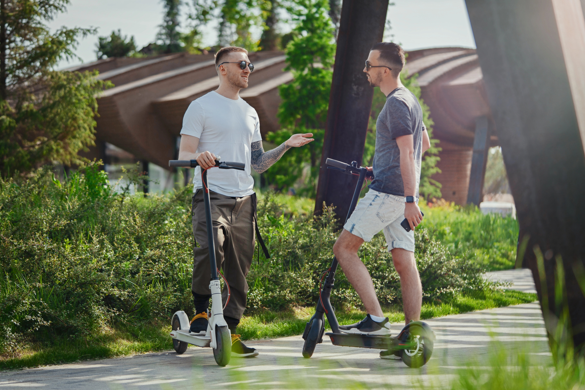 Two people standing with their electric scooters on a paved path in a park. One wears a white shirt and dark pants, the other wears a gray shirt and shorts. They are engaged in conversation, with greenery and a modern structure in the background.