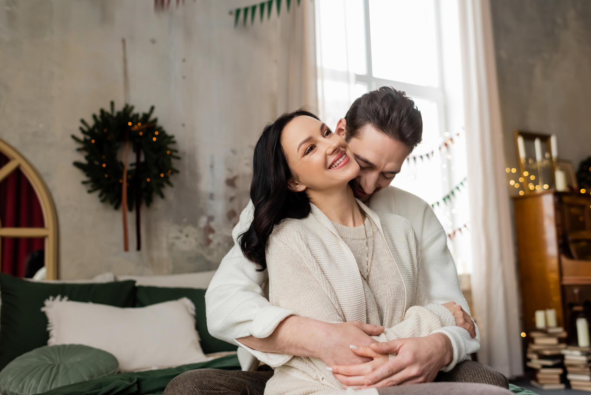 A couple is sitting together in a cozy room decorated for the holidays. The woman, wearing a light-colored sweater, is smiling and leaning back against the man, who is hugging her from behind. There is a wreath on the wall and holiday decorations in the background.