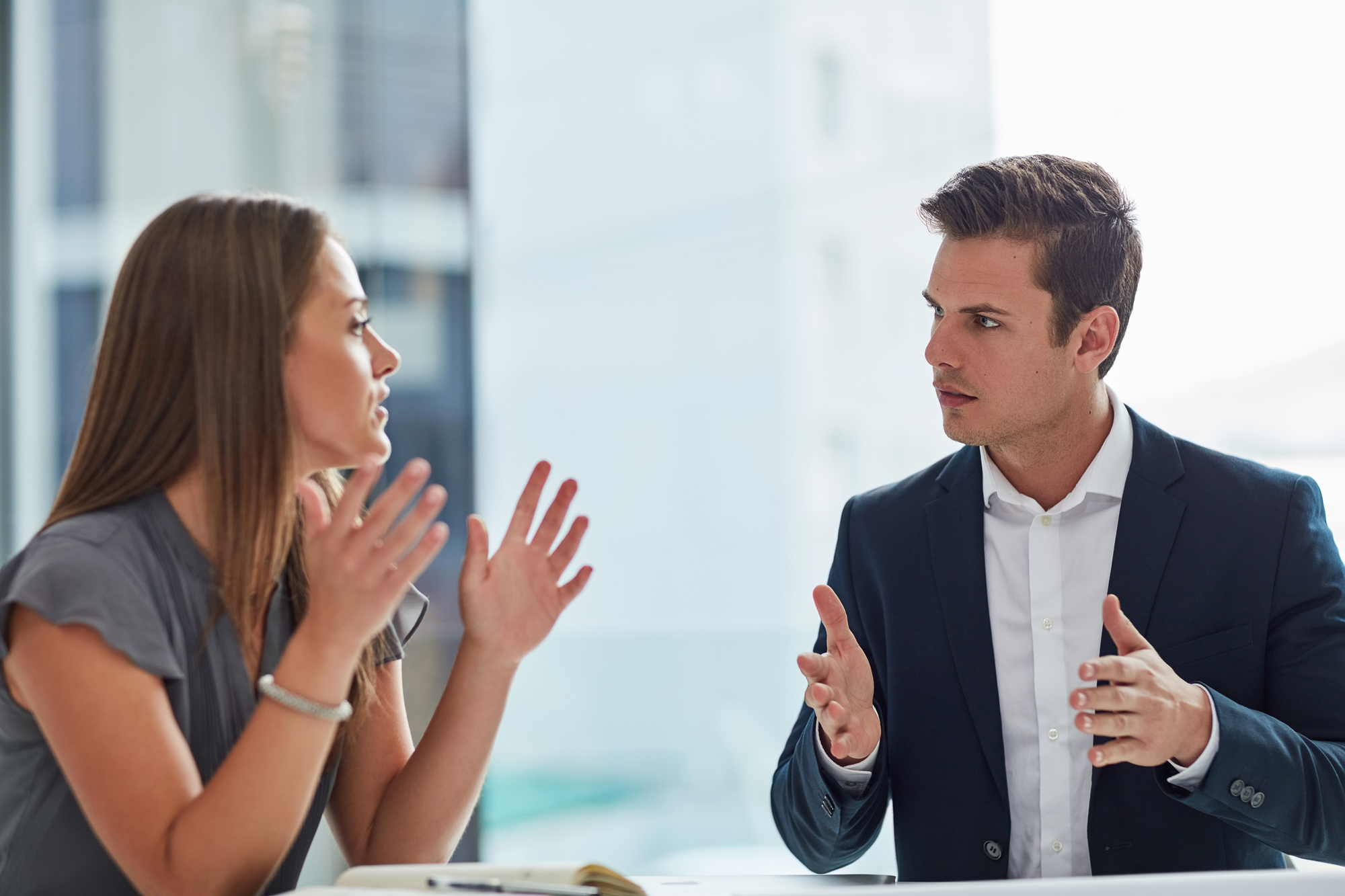 A woman and man are engaged in a serious conversation in an office setting. The woman, on the left, is gesturing with her hands, while the man, on the right, is responding with a focused expression and hand gestures. Both are dressed in business attire.