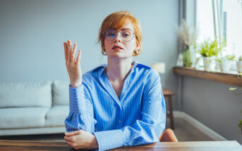 A person with red hair and glasses sits at a wooden table in a modern home interior. They are wearing a blue button-up shirt and have a contemplative expression, with one hand raised and the other resting on the table. The background includes a couch and various plants.