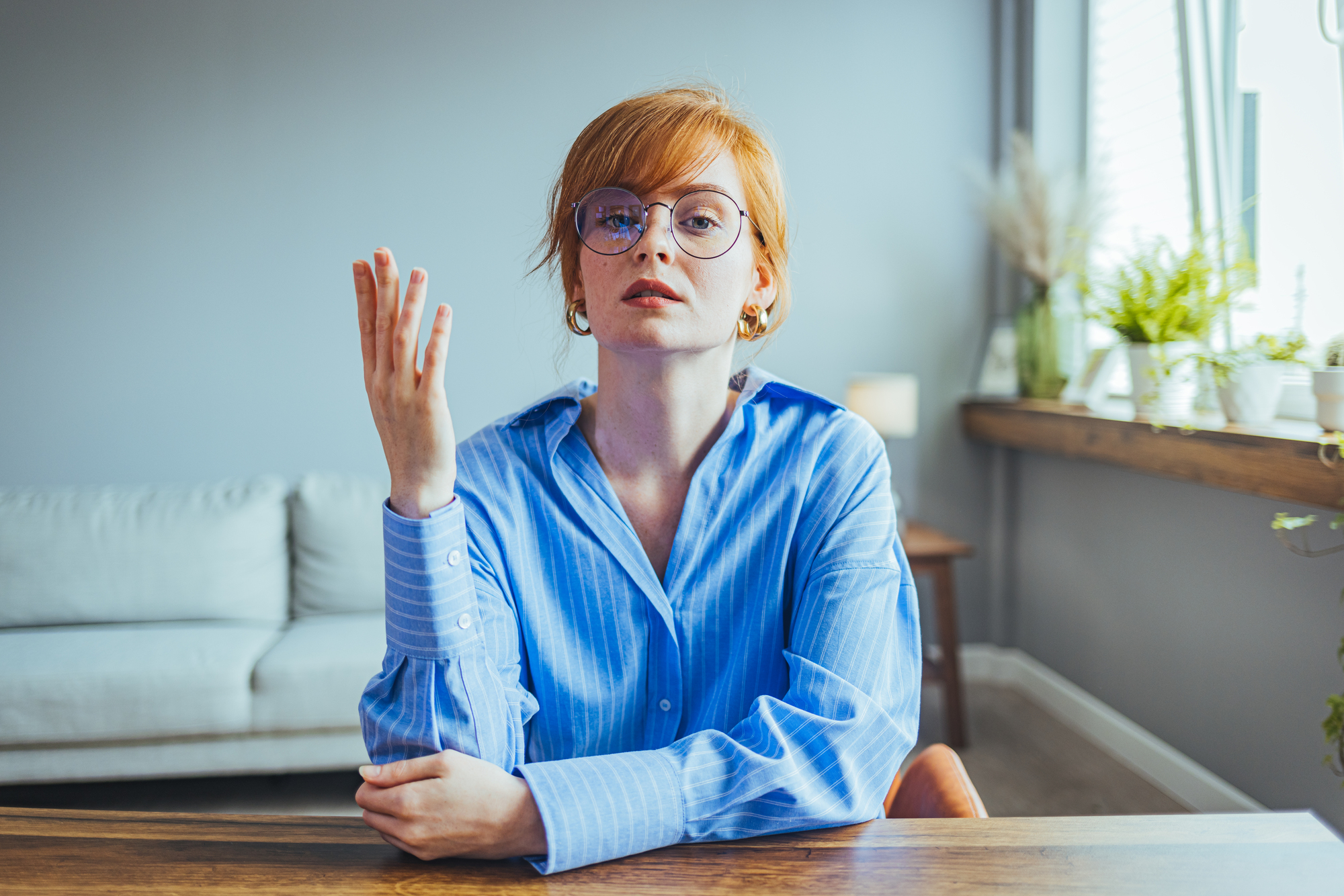 A person with red hair and glasses sits at a wooden table in a modern home interior. They are wearing a blue button-up shirt and have a contemplative expression, with one hand raised and the other resting on the table. The background includes a couch and various plants.