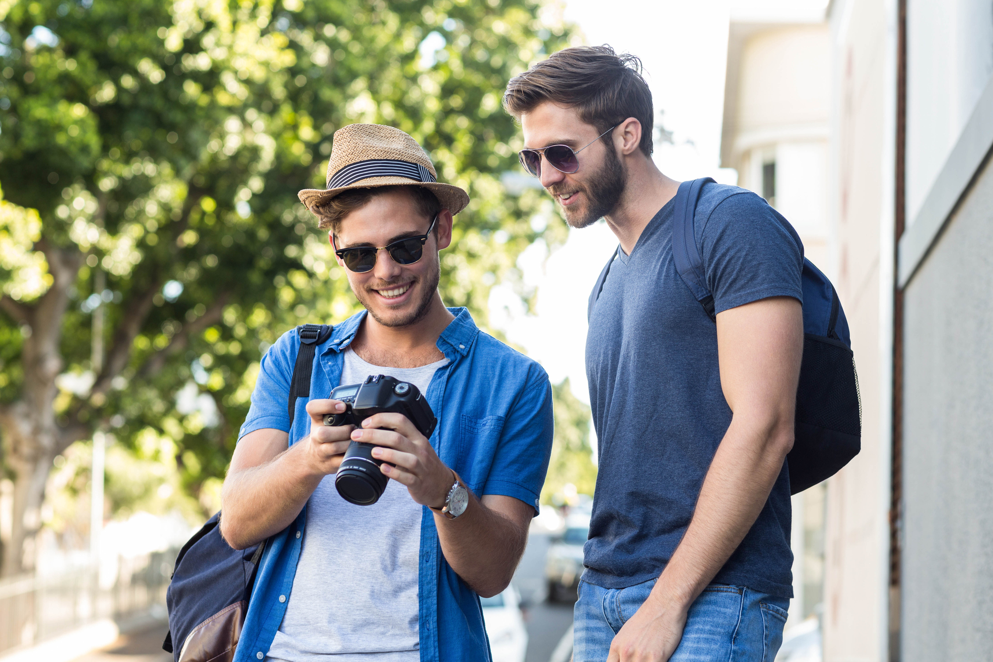 Two young men stand outdoors on a sunny day. One, wearing a straw hat, sunglasses, and a blue shirt, holds a camera and shows the screen to the other, who is wearing a gray T-shirt, sunglasses, and a backpack. They appear to be reviewing photos and smiling.