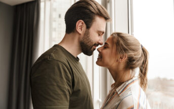 A smiling couple stands close together by a window, with their foreheads touching. The man has a beard and is wearing a green shirt, while the woman has her hair in a ponytail and is dressed in a striped shirt. Natural light illuminates the room.