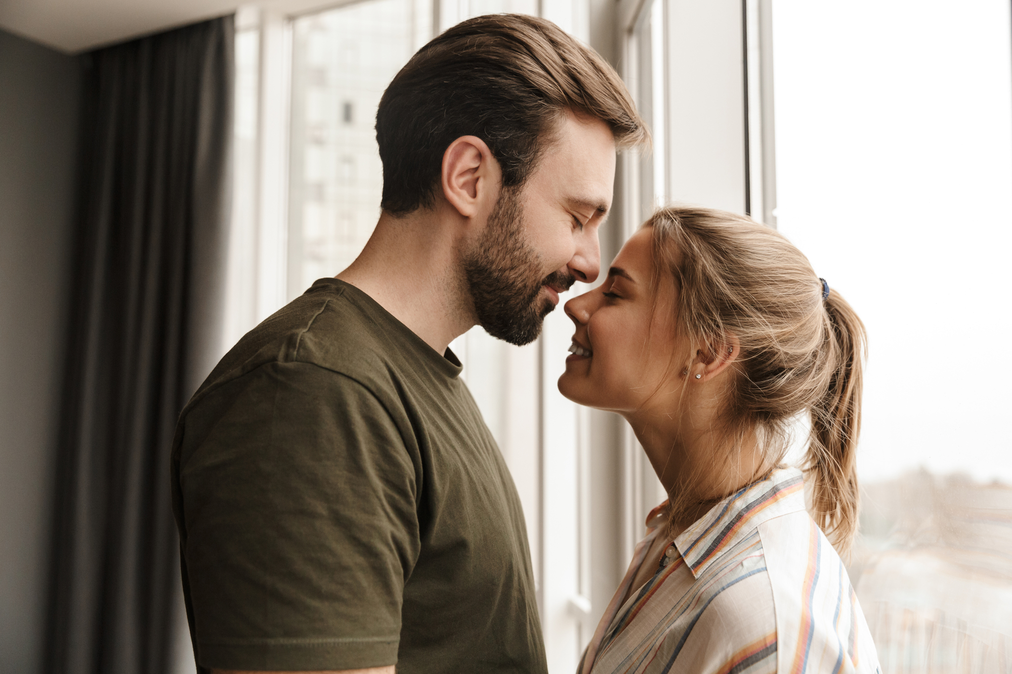 A smiling couple stands close together by a window, with their foreheads touching. The man has a beard and is wearing a green shirt, while the woman has her hair in a ponytail and is dressed in a striped shirt. Natural light illuminates the room.